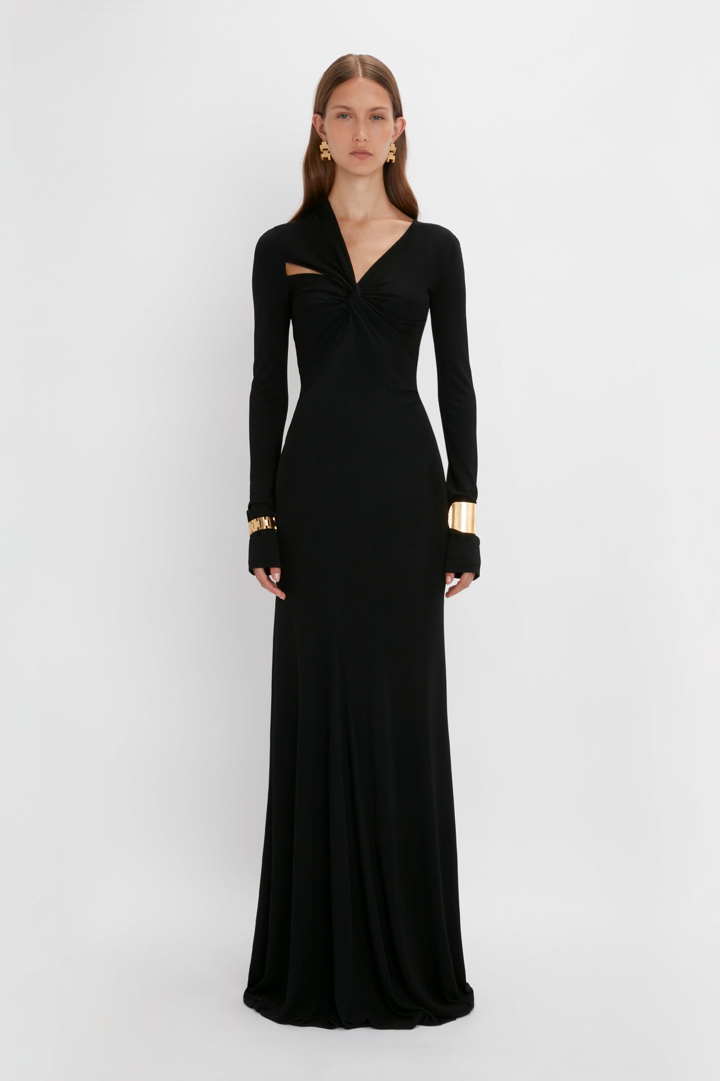 A woman in a long-sleeve black gown with a twist-front design, accessorized with contemporary accessories including Victoria Beckham's Exclusive Perfume Cuff In Gold earrings and bracelets, standing against a white background.
