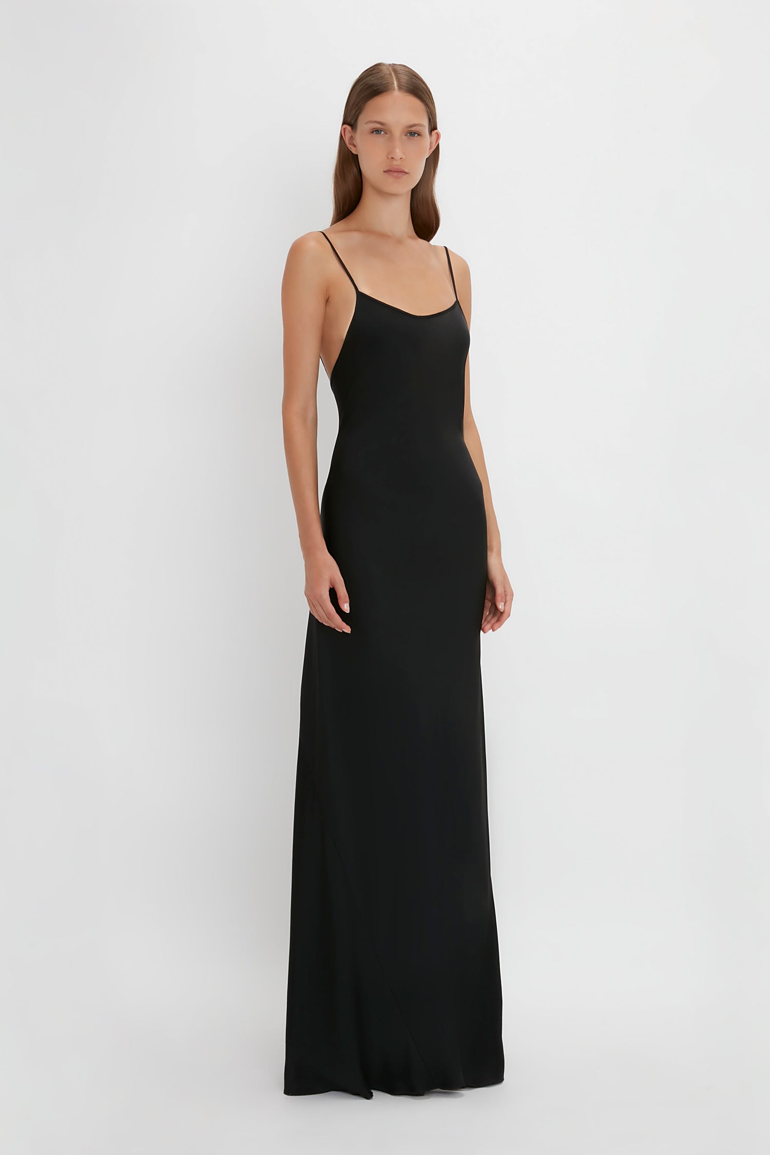 A woman stands against a white background wearing the Victoria Beckham Floor-Length Cami Dress In Black, evoking the elegance of 90s fashion.