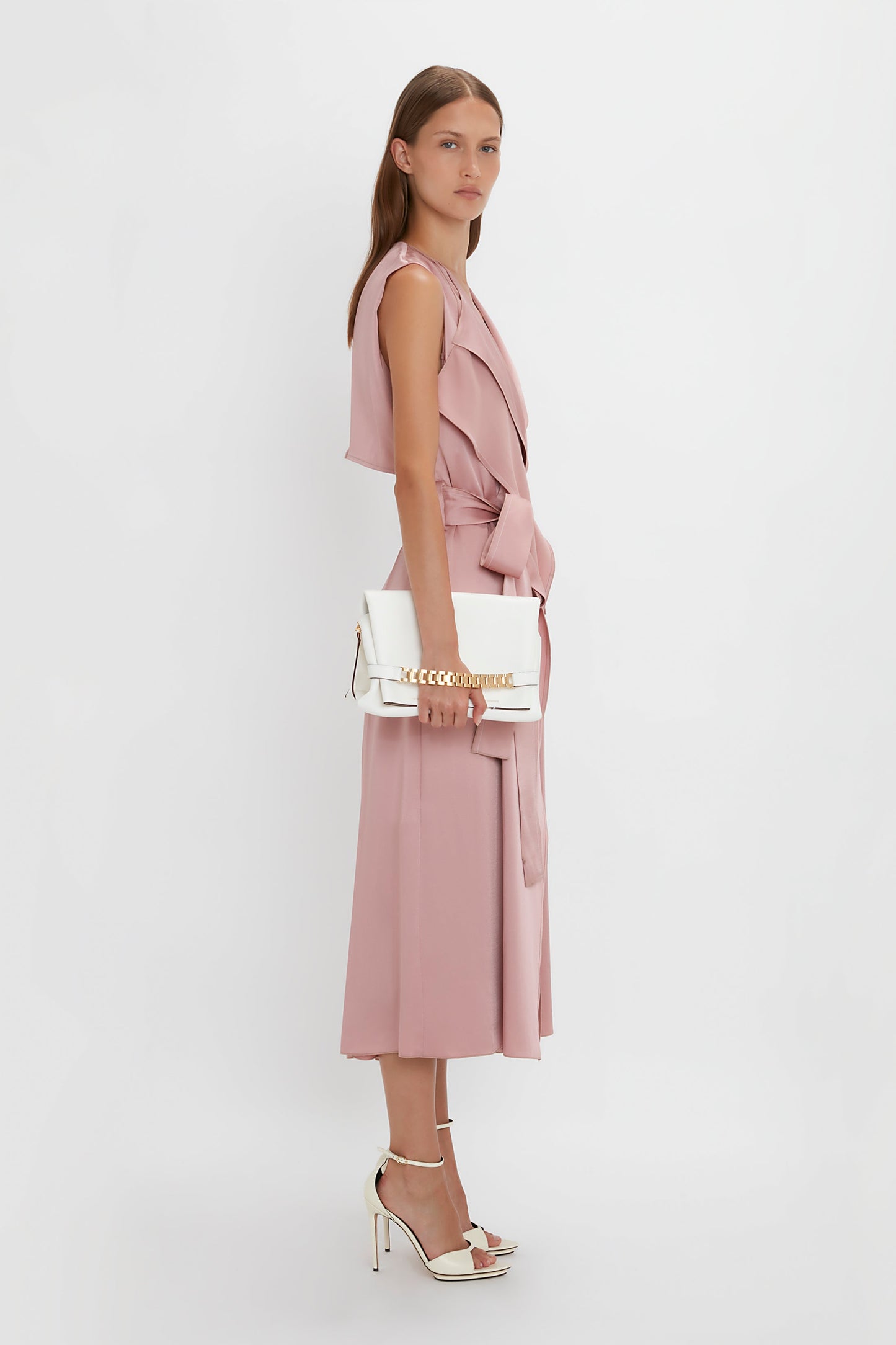 A person in a pink sleeveless dress and white high-heeled sandals holds a Victoria Beckham Chain Pouch Bag With Strap In White Leather against a plain white background.