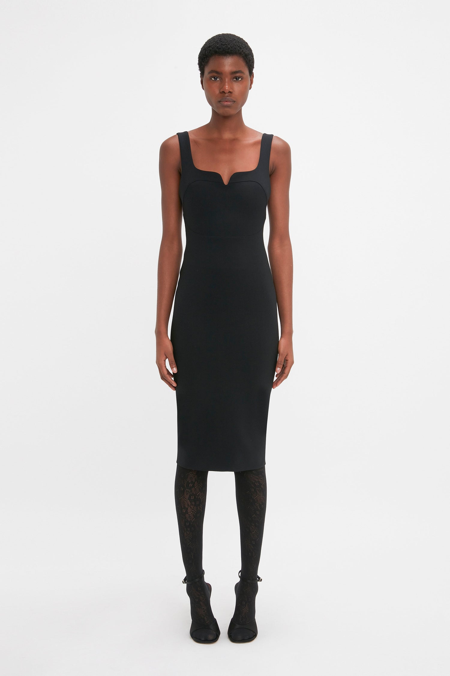 Person standing against a plain white background, wearing a Sleeveless Fitted T-Shirt Dress In Black by Victoria Beckham, patterned black tights, and black shoes. The expression is neutral.