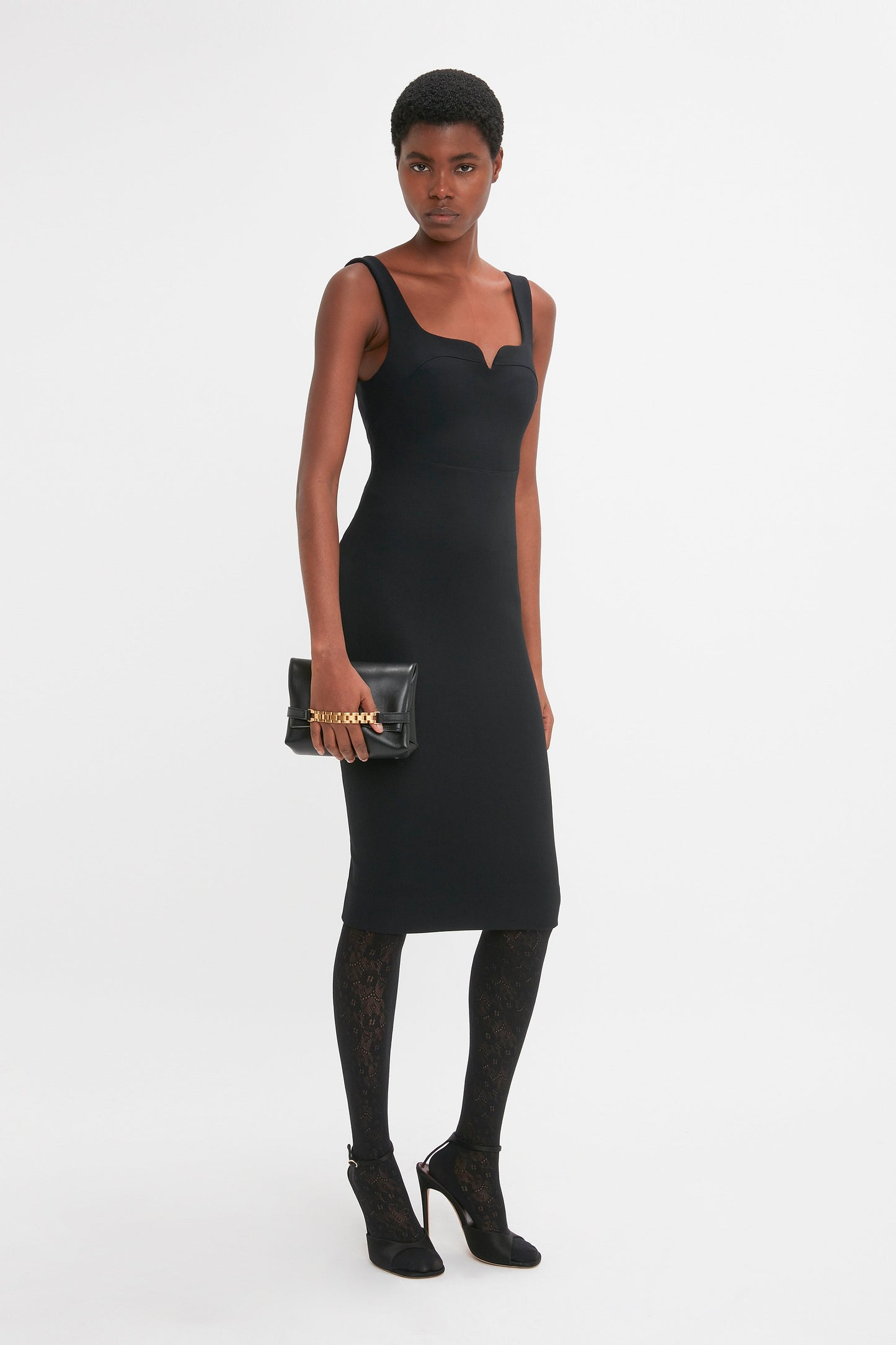 Person standing against a white background, wearing the Victoria Beckham Sleeveless Fitted T-Shirt Dress In Black, black patterned tights, and black high-heeled shoes. They are holding a black clutch bag.