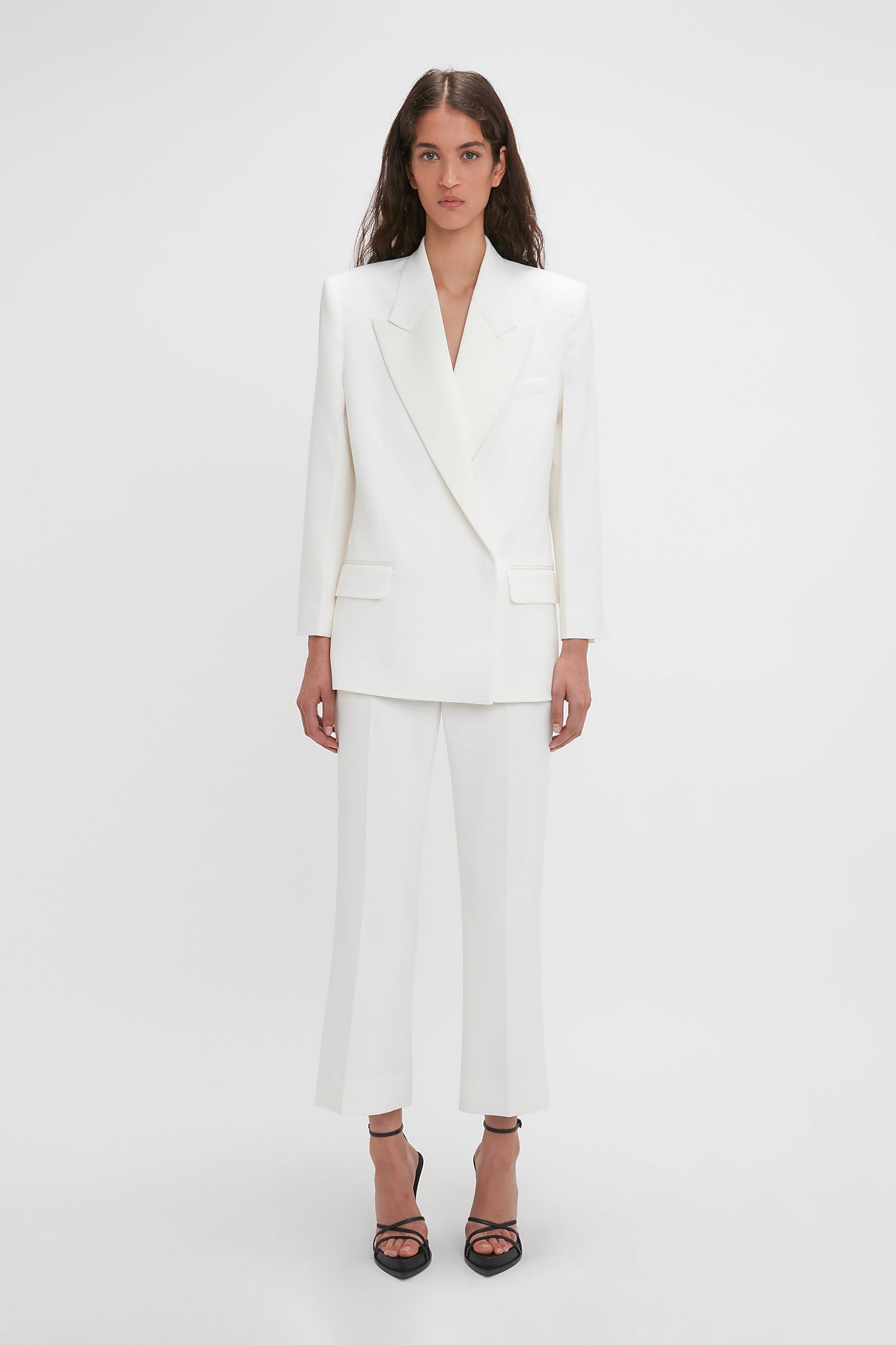 A woman stands against a plain background, wearing the Victoria Beckham Exclusive Double Breasted Tuxedo Jacket In Ivory with satin peak lapels and matching white trousers, paired with black-heeled sandals.