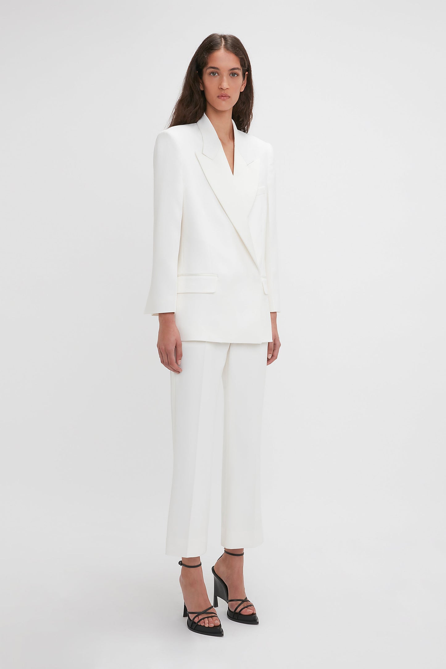 A woman is standing against a white background, wearing the Victoria Beckham Exclusive Double Breasted Tuxedo Jacket In Ivory with satin peak lapels and black high-heeled sandals.