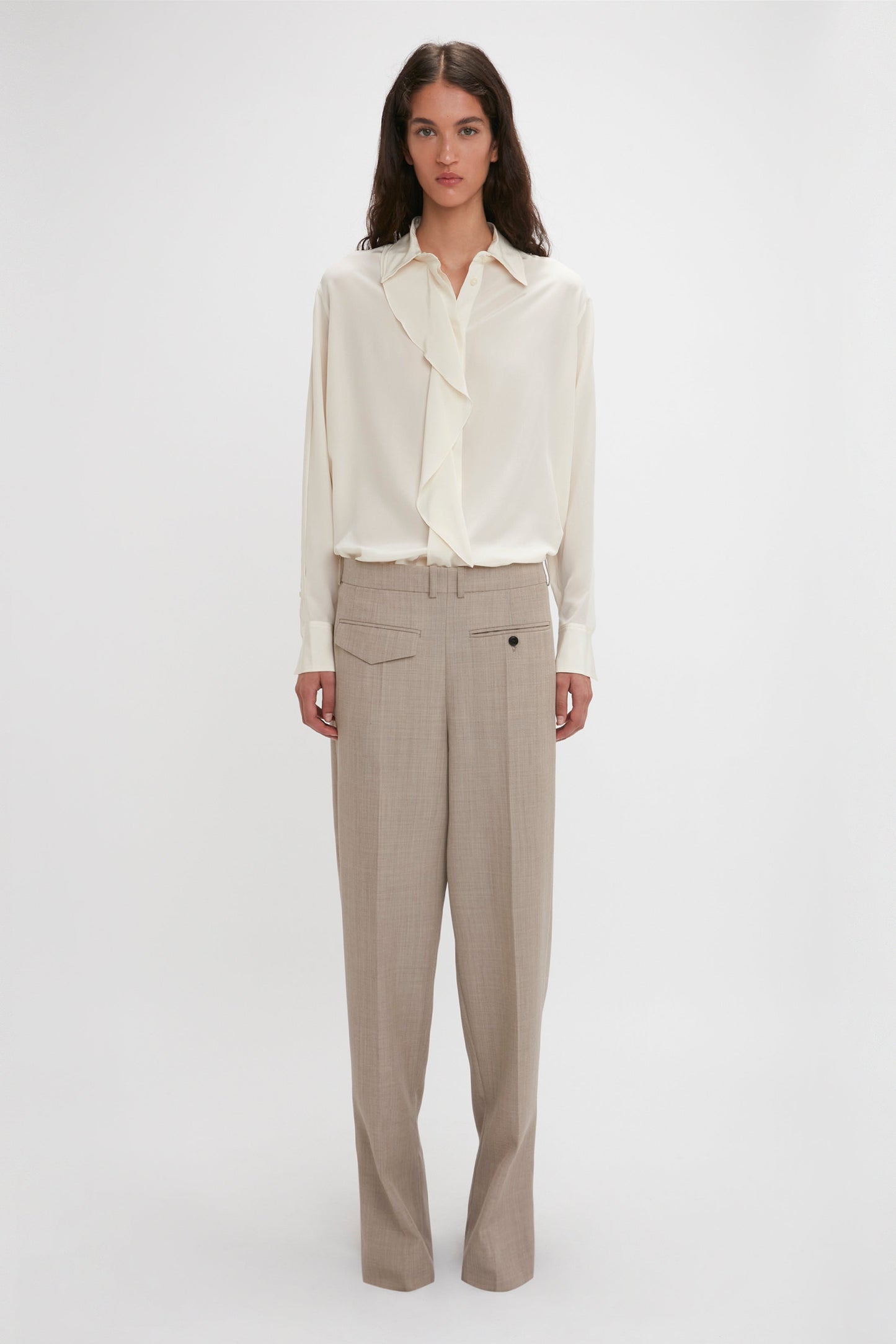 A person stands against a plain background wearing an Asymmetric Ruffle Blouse In Ivory by Victoria Beckham, featuring voluminous sleeves, and beige trousers.