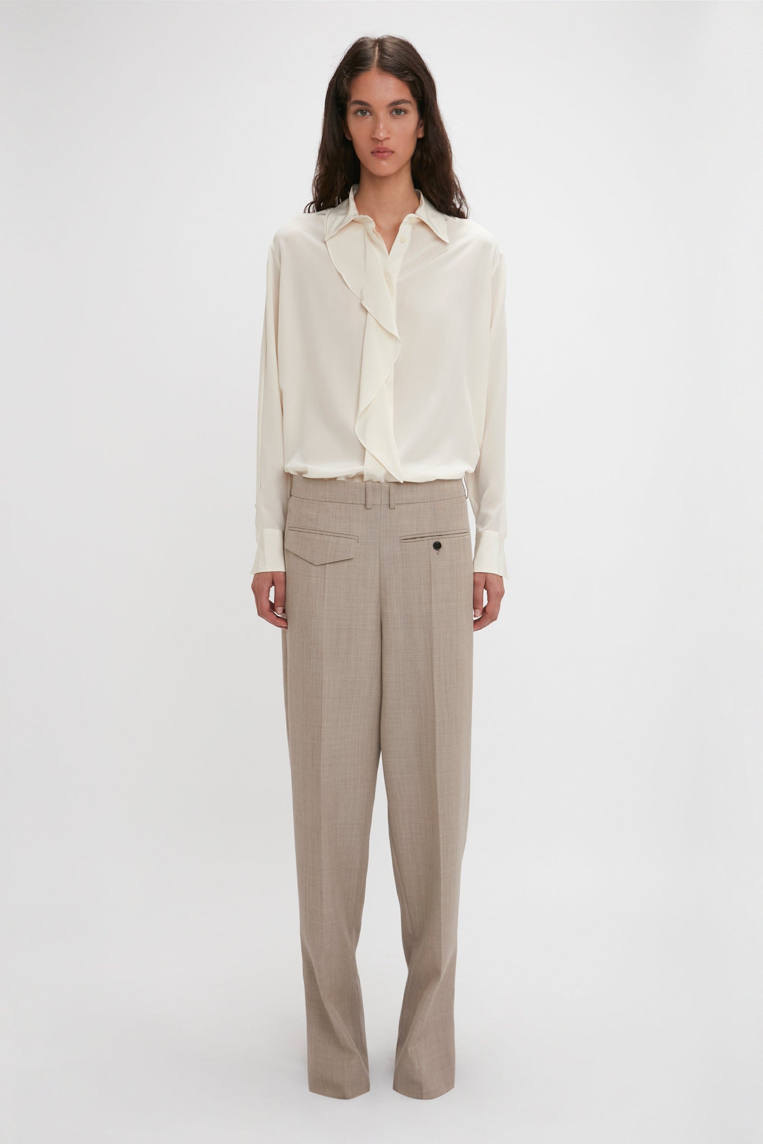 A person stands against a plain background wearing an Asymmetric Ruffle Blouse In Ivory by Victoria Beckham, featuring voluminous sleeves, and beige trousers.