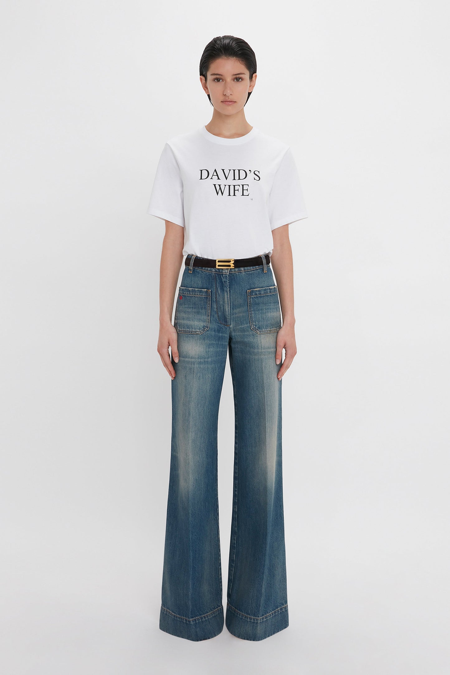 A woman wearing a white Victoria Beckham 'David's Wife' Slogan T-Shirt with "David's Wife" text and blue high-waisted, wide-leg jeans, paired with a black belt.