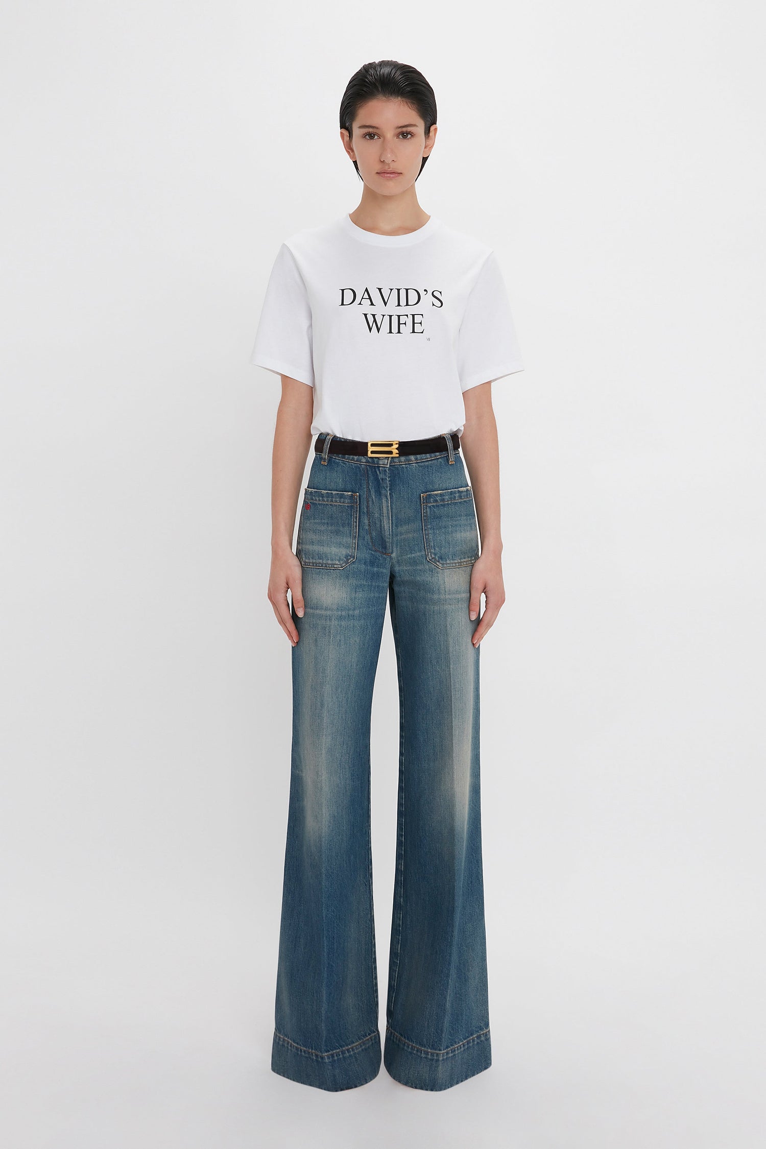 Person wearing a white T-shirt with "DAVID'S WIFE" written on it, paired with Victoria Beckham's Alina High Waisted Jean In Indigrey Wash and a black belt.