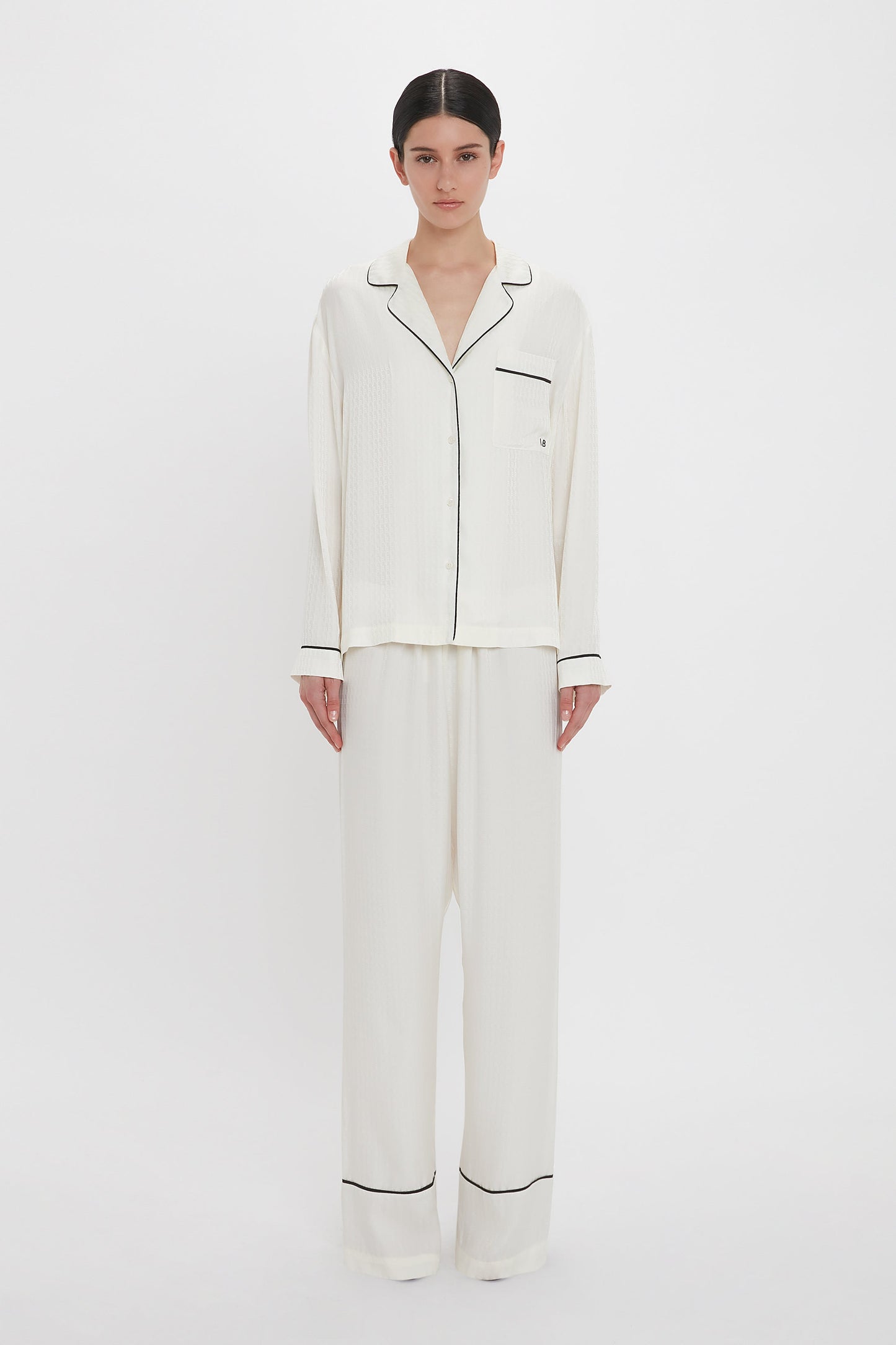 A person with dark hair stands against a white background wearing the Victoria Beckham VB Monogram Pyjama Sleep Set In Ivory.