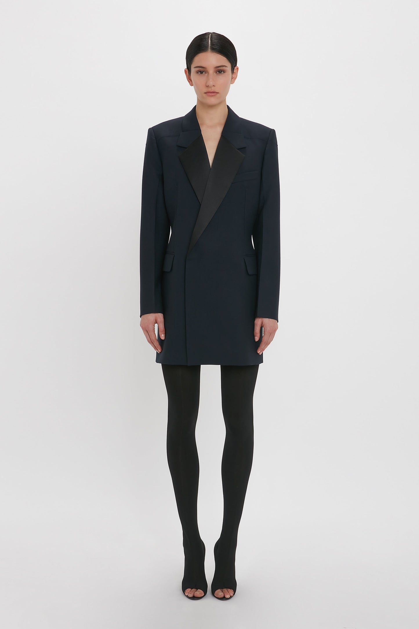 Person in an Exclusive Fold Shoulder Detail Dress In Midnight by Victoria Beckham, worn over black tights and open-toed shoes, standing against a plain white background.