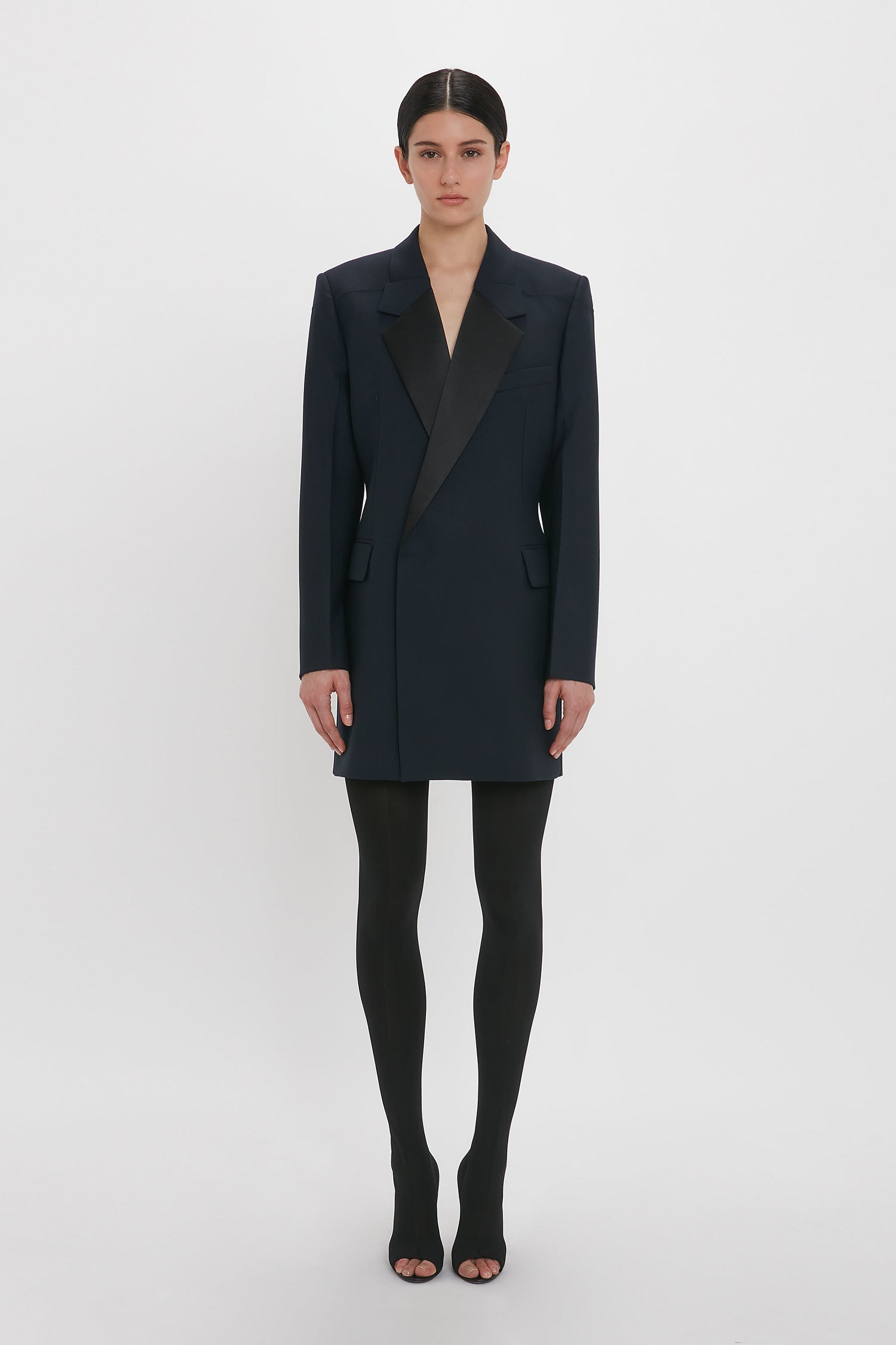 Person in an Exclusive Fold Shoulder Detail Dress In Midnight by Victoria Beckham, worn over black tights and open-toed shoes, standing against a plain white background.