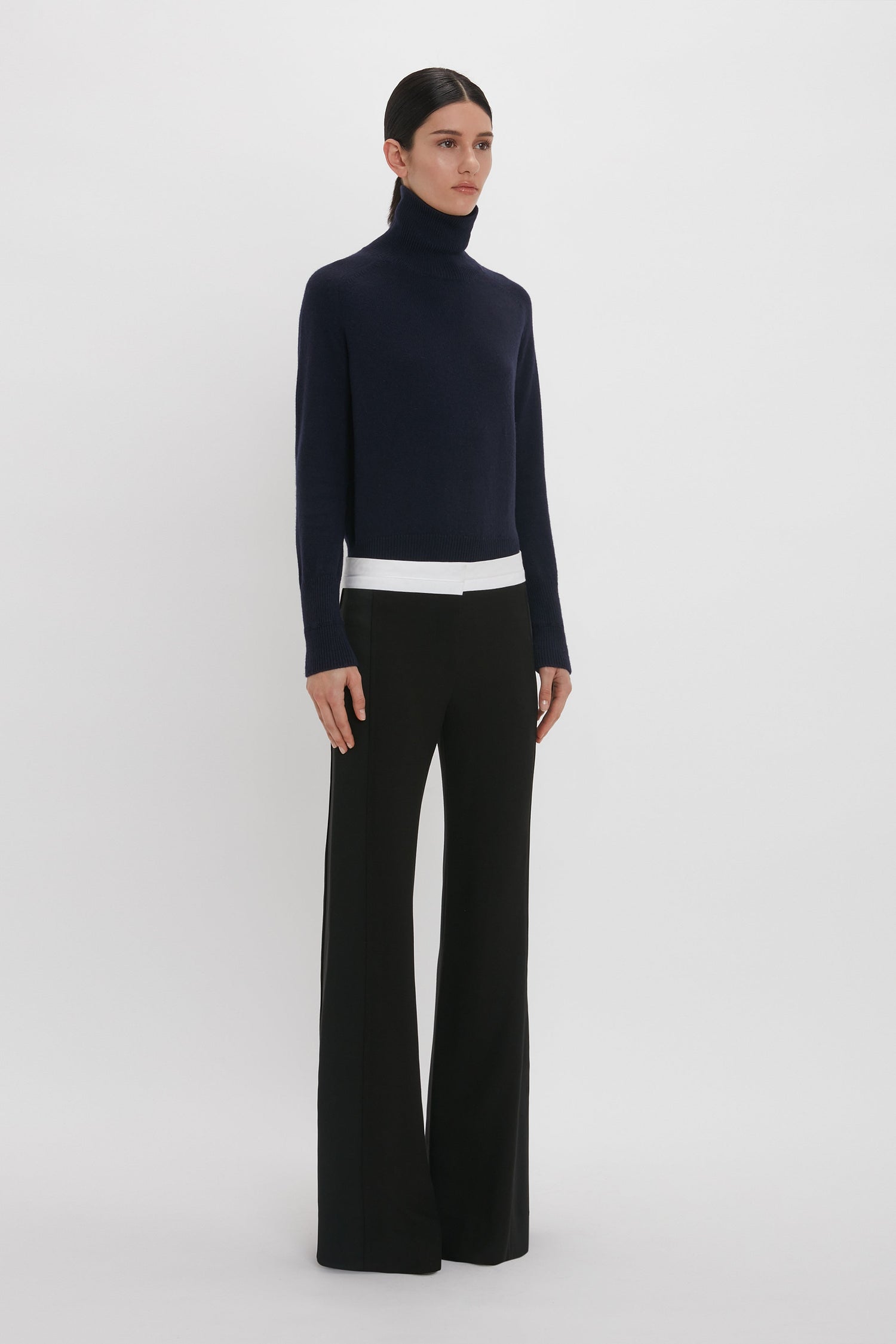 Person standing against a plain background, wearing a dark turtleneck sweater and Victoria Beckham Side Panel Trouser In Black.