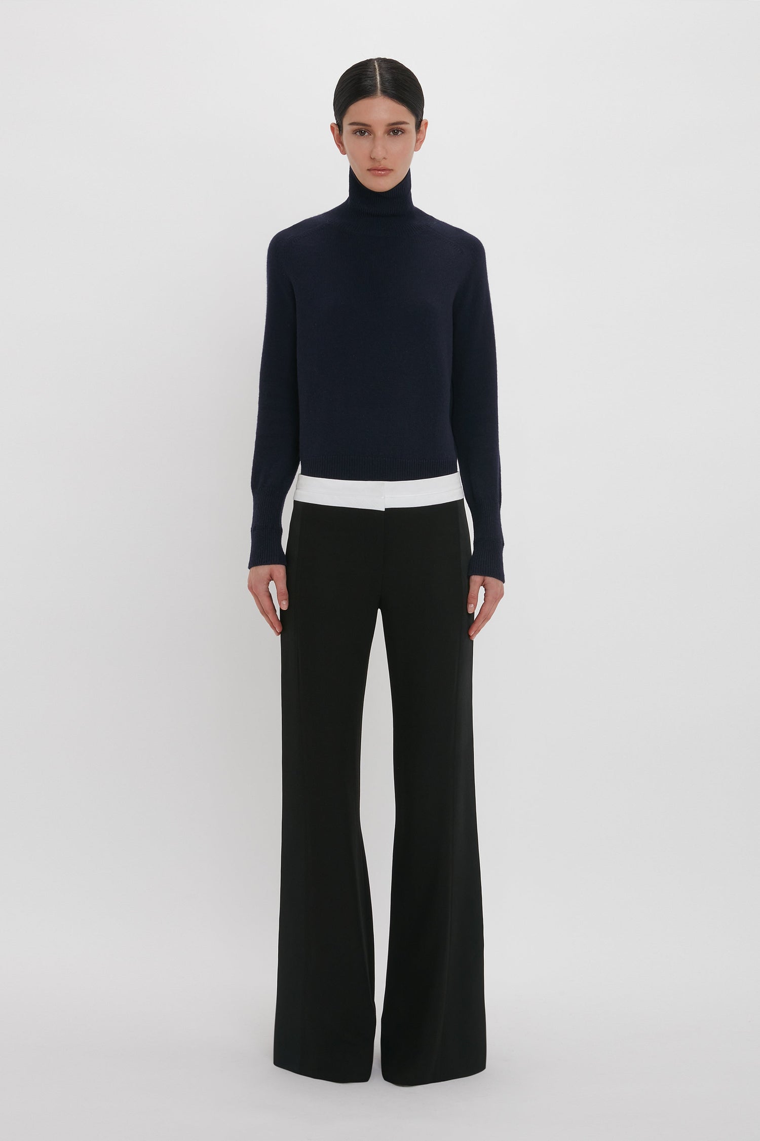 A person stands against a plain white background, wearing a dark navy turtleneck sweater, white undershirt, and the Side Panel Trouser In Black by Victoria Beckham with a wide-leg design. They have dark hair tied back.