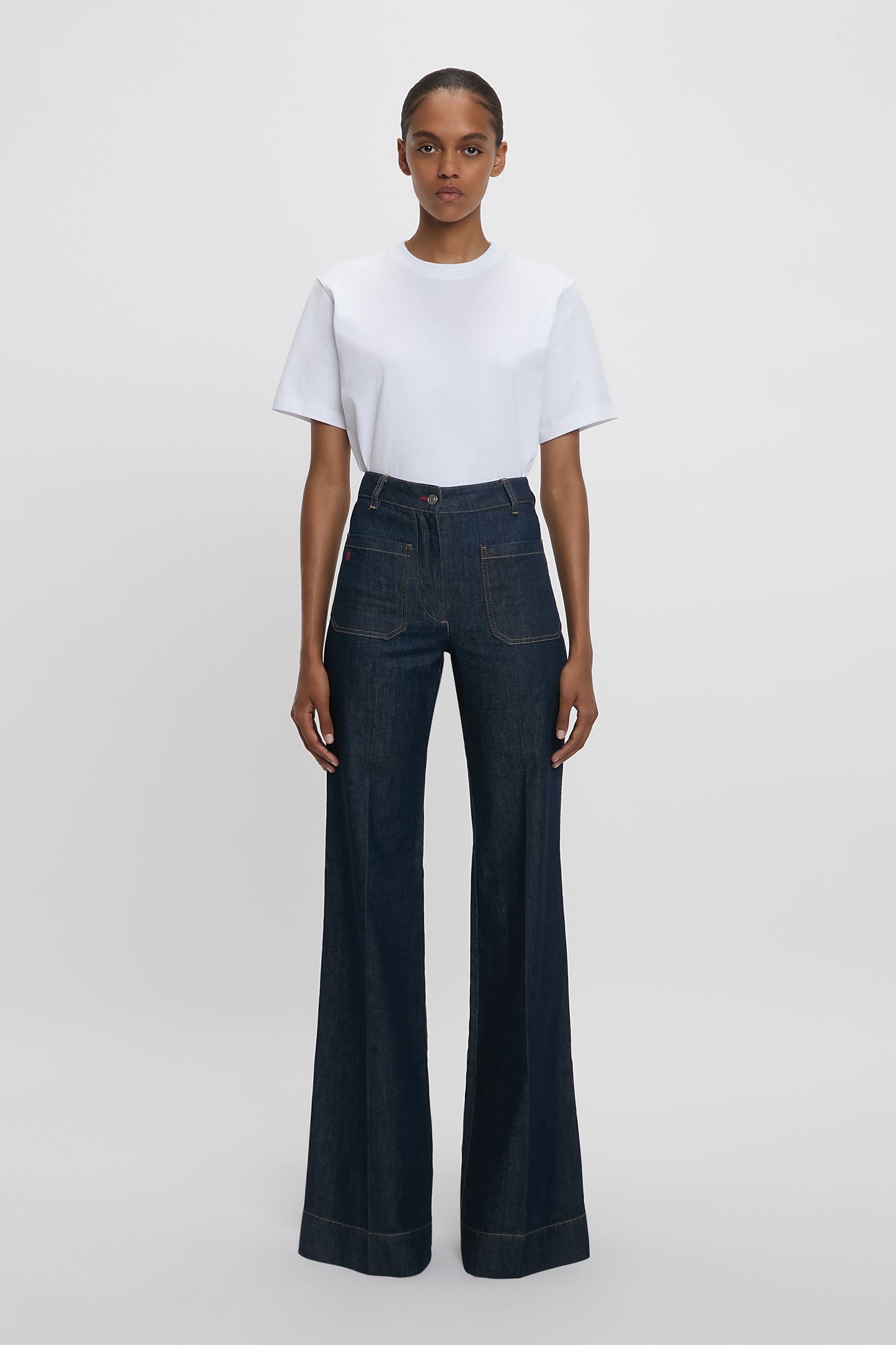 A woman standing against a white background wearing a white t-shirt and high-waisted, flared, indigo blue denim jeans with a branded leather patch at the waistband.