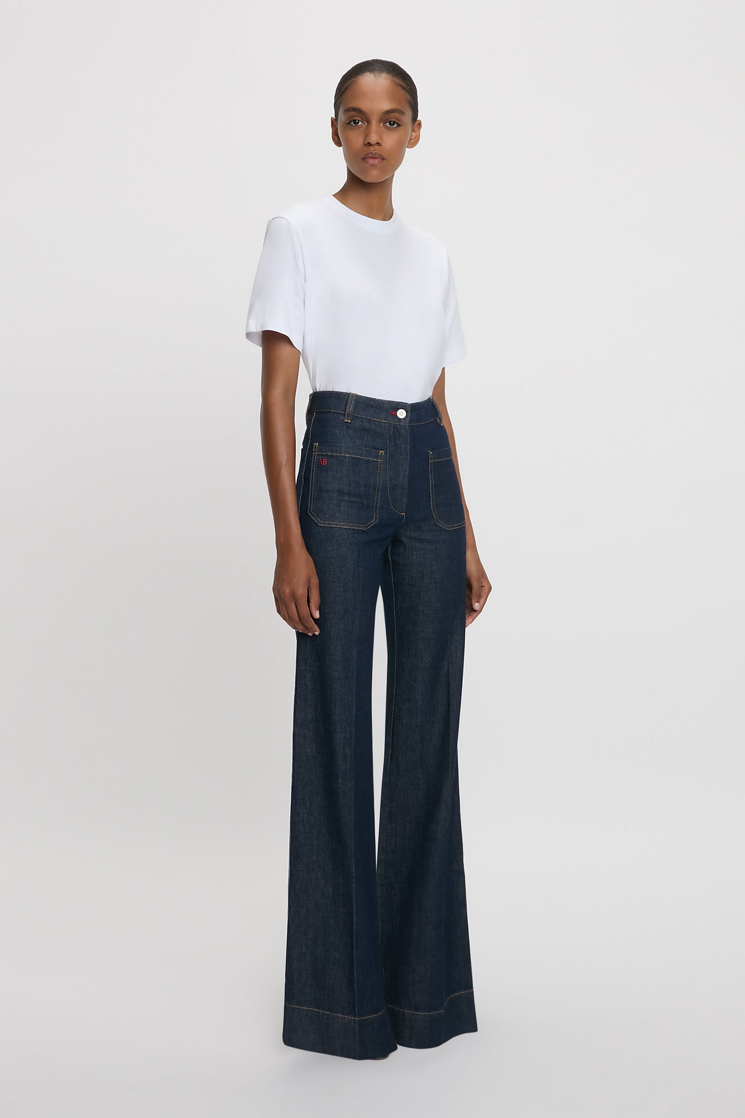 A woman standing at an angle against a white background wearing a white t-shirt and high-waisted, flared, indigo blue denim jeans with a branded leather patch at the waistband.