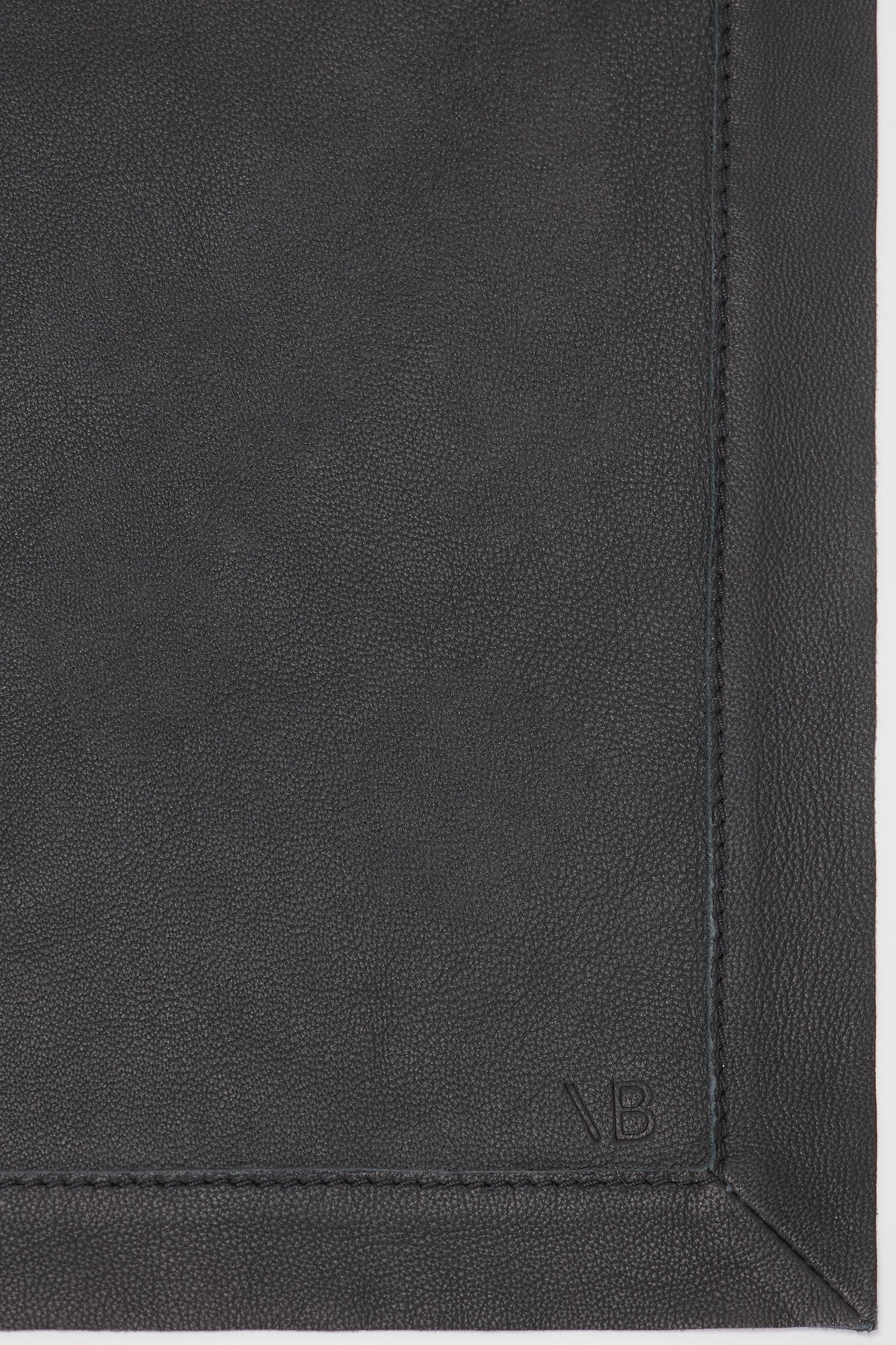 Close-up of a dark, textured leather scarf with visible stitching and the initials "VB" embossed at the bottom right corner, showcasing its classic black hue and supple finish. The product is the Victoria Beckham Leather Scarf In Black.