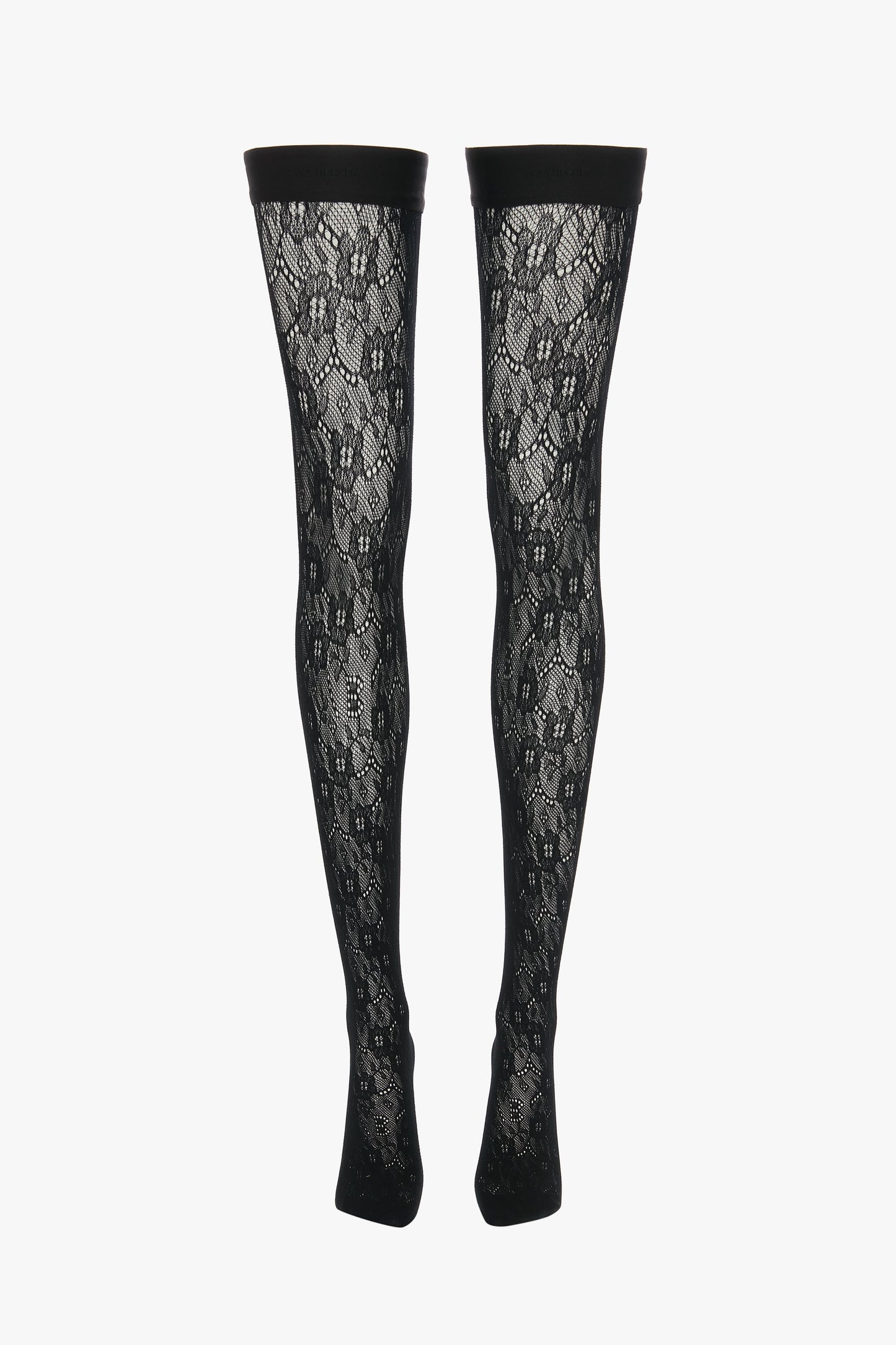 A pair of Exclusive VB Monogram Lace Hold-Ups In Black by Victoria Beckham, these black lace patterned thigh-high stockings feature a sheer finish and a solid black band at the top, displayed vertically.