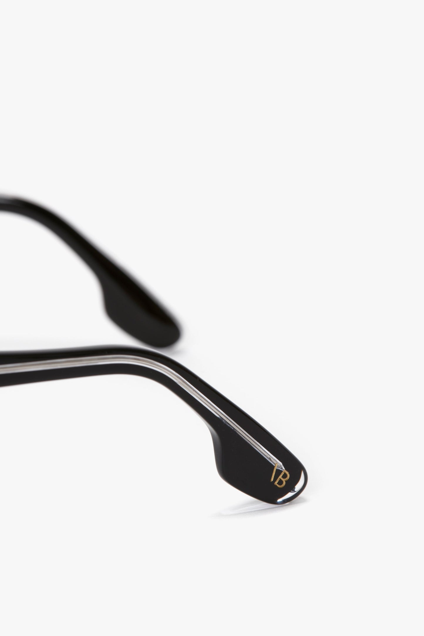 Close-up of rectangular-shaped black glasses frames showing the acetate temples with a small gold "VB" logo near the edges. The background is plain white. The product featured is Layered Mask Sunglasses In Black-Silver-Mirror by Victoria Beckham.
