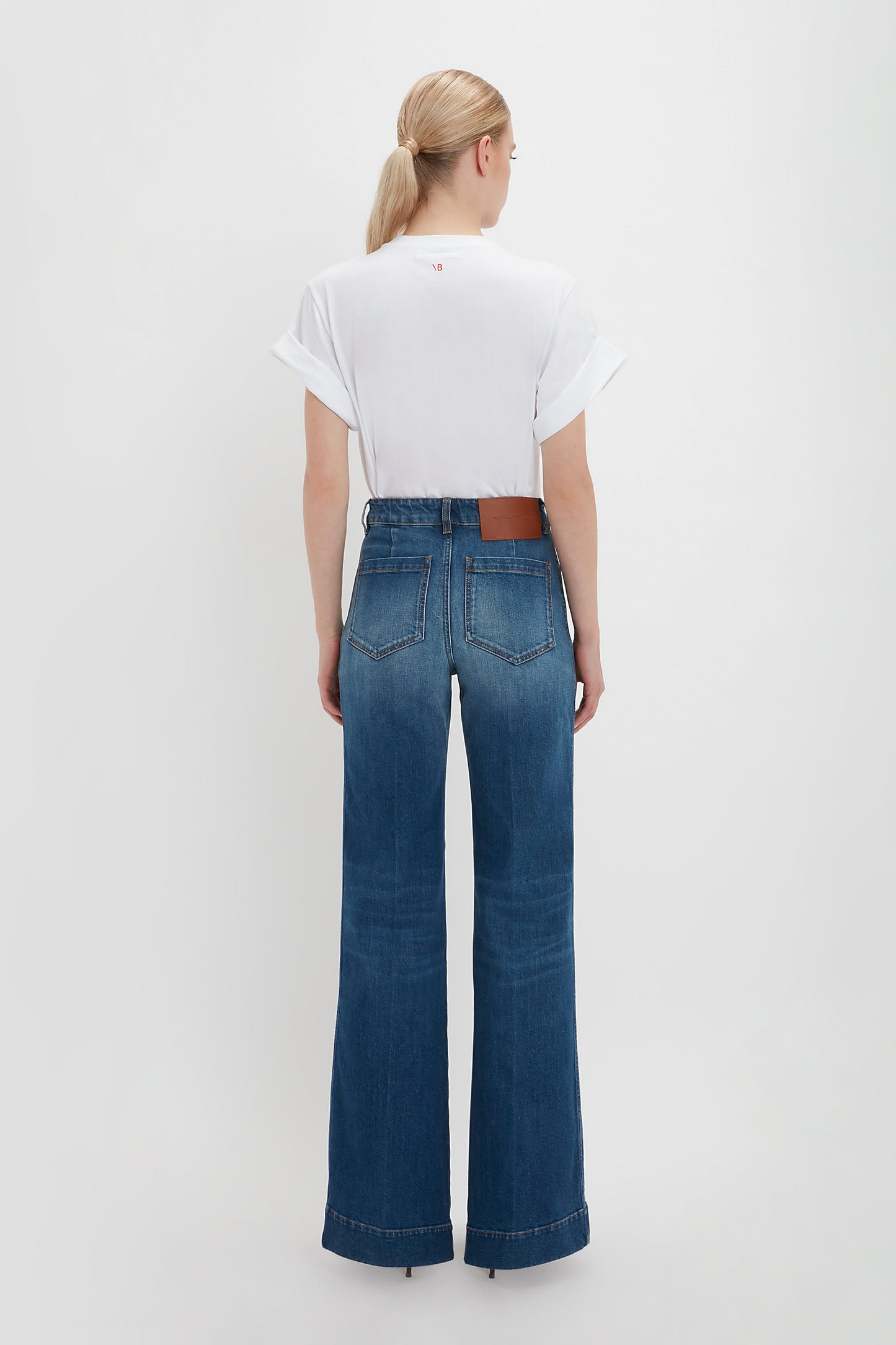 A person with blond hair in a ponytail, wearing an oversized Victoria Beckham Asymmetric Relaxed Fit T-Shirt In White and blue high-waisted wide-leg jeans with asymmetric folded sleeves, stands facing away.