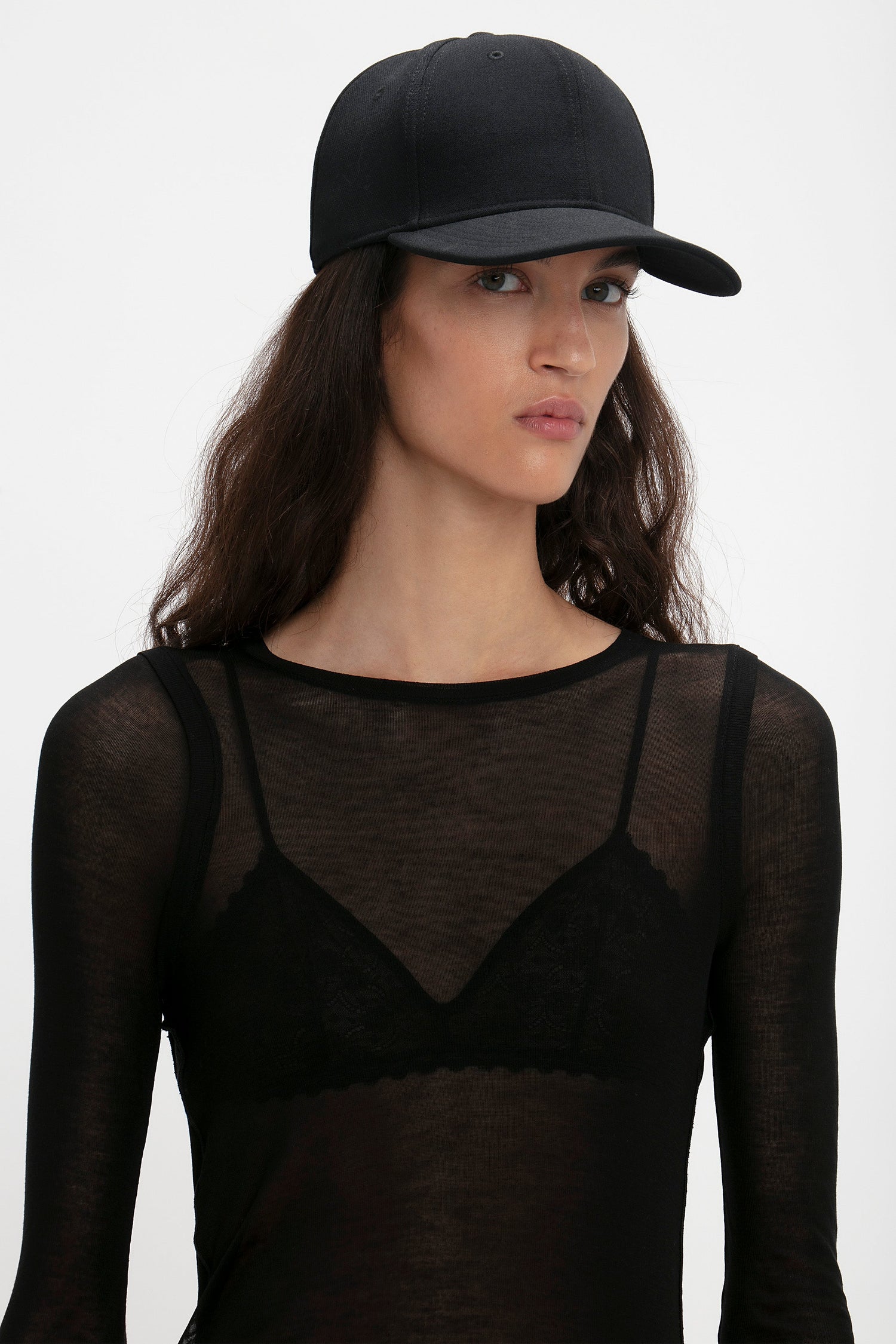 Woman wearing a black Exclusive Logo Cap In Black by Victoria Beckham and sheer top with lace detailing, looking to the side against a white background.