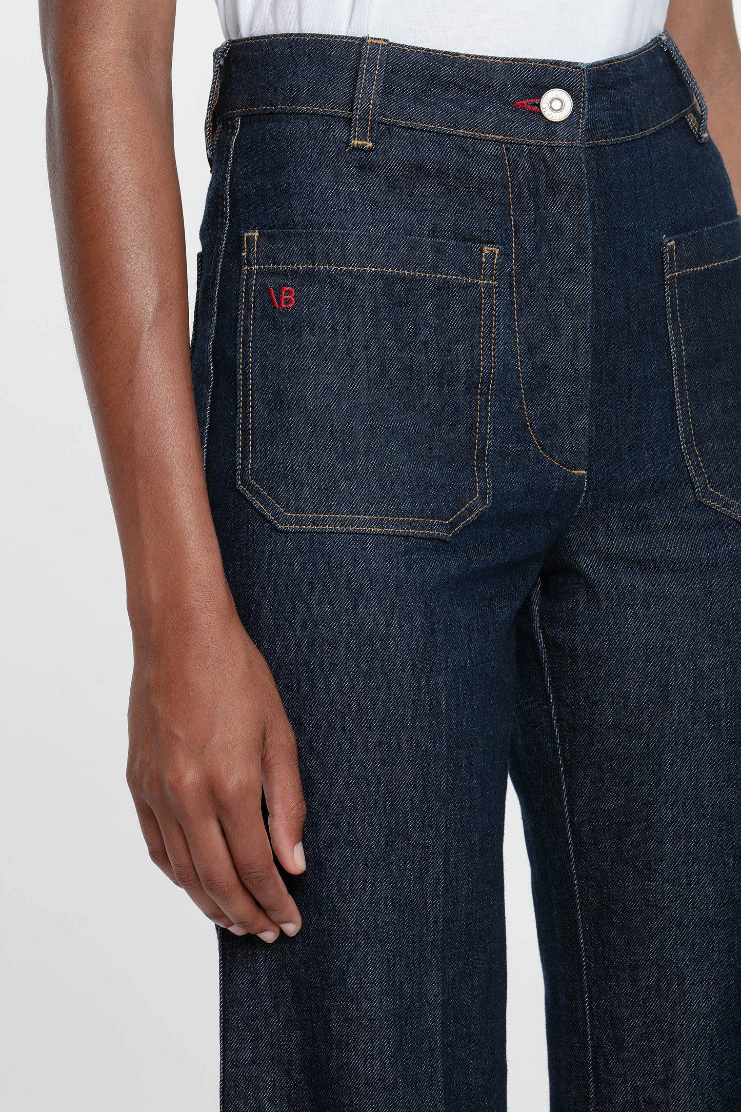 A woman standing against a white background wearing a white t-shirt and high-waisted, flared, indigo blue denim jeans with a branded leather patch at the waistband.