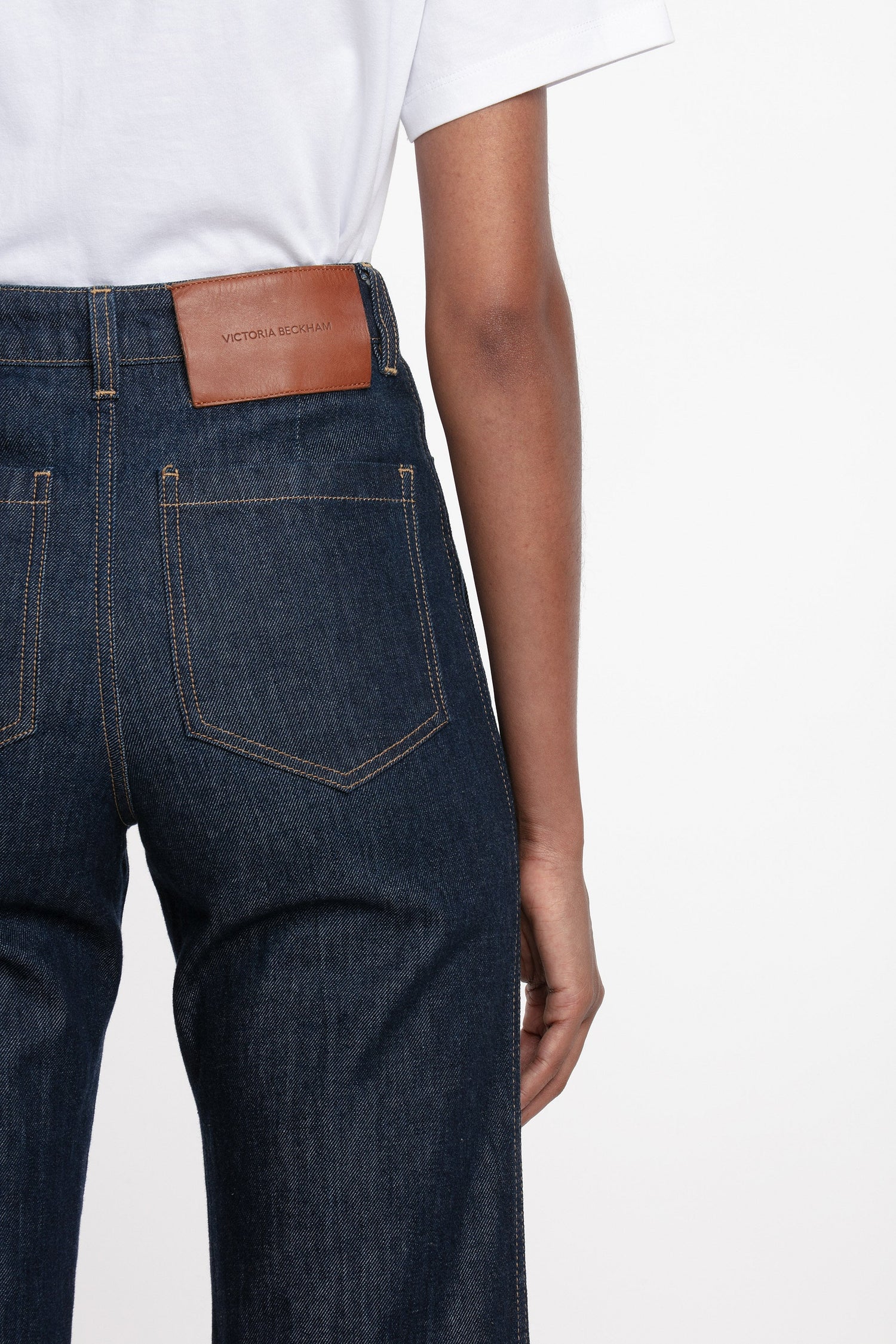 A woman standing against a white background wearing a white t-shirt and high-waisted, flared, indigo blue denim jeans with a branded leather patch at the waistband.