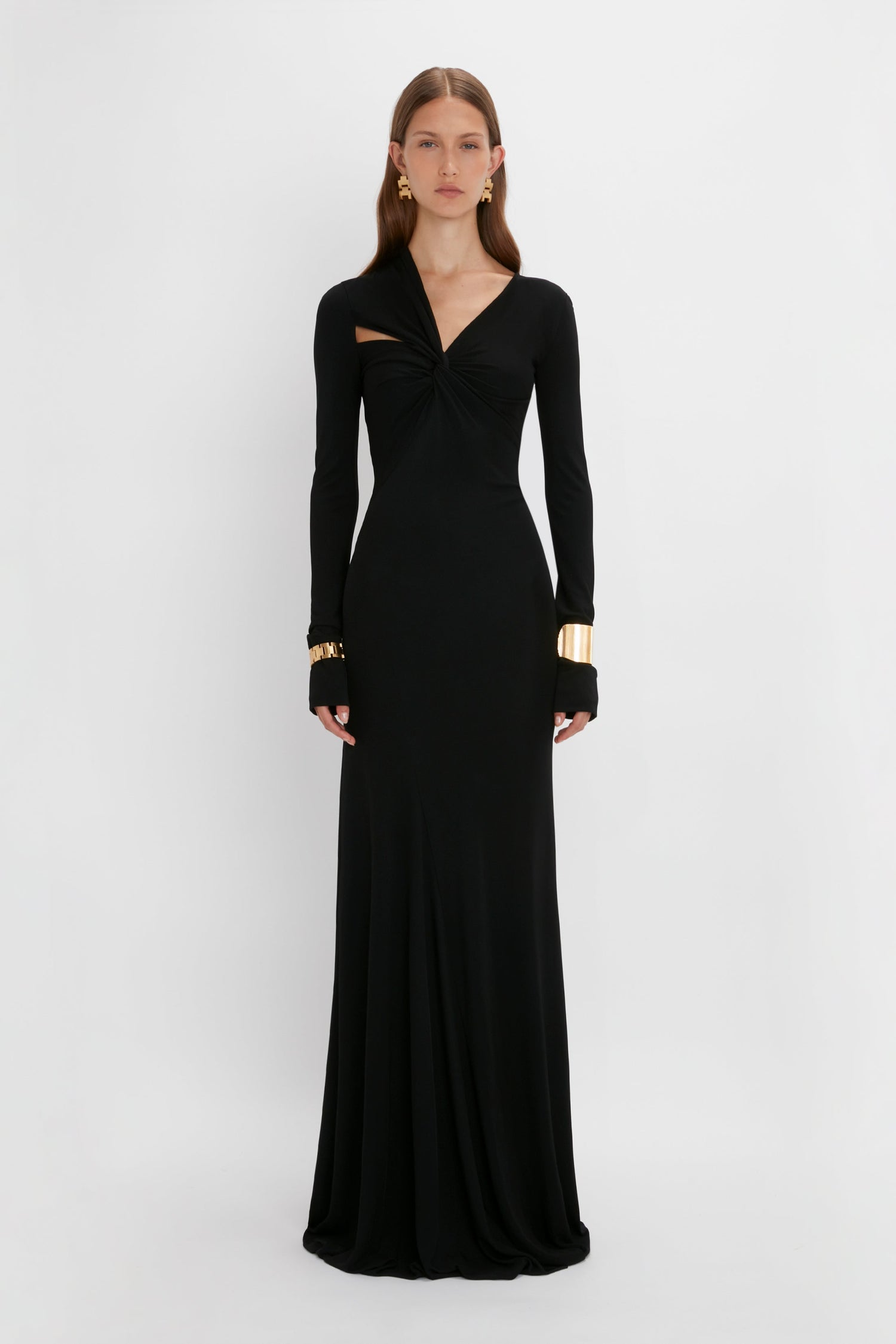 A woman in a long-sleeve black gown with a twist-front design, accessorized with contemporary accessories including Victoria Beckham's Exclusive Perfume Cuff In Gold earrings and bracelets, standing against a white background.