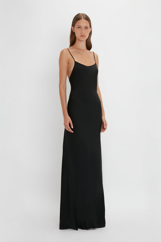 A woman stands against a white background wearing the Victoria Beckham Floor-Length Cami Dress In Black, evoking the elegance of 90s fashion.