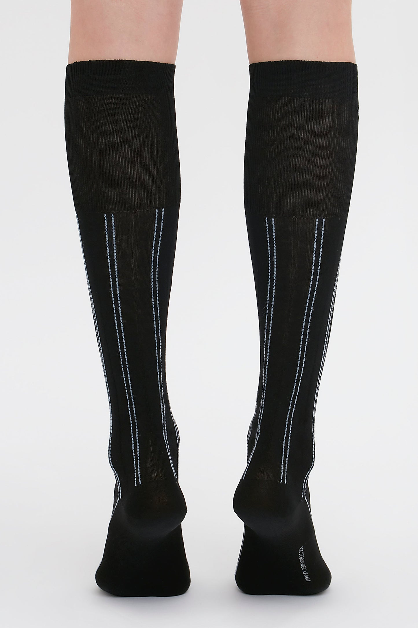 A person wearing black Superfine Rib Socks In Black by Victoria Beckham with thin blue and white vertical stripes on the back, standing on a white background.