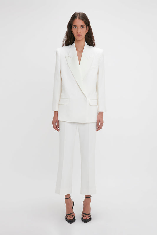A woman stands against a plain background, wearing the Victoria Beckham Exclusive Double Breasted Tuxedo Jacket In Ivory with satin peak lapels and matching white trousers, paired with black-heeled sandals.
