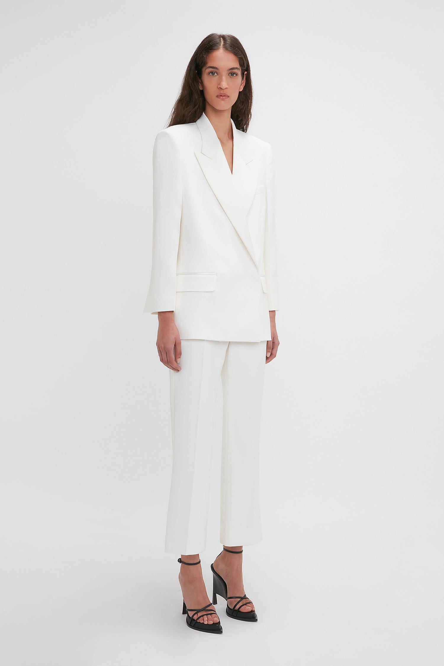 A woman stands facing forward, wearing a white suit featuring a double-breasted blazer and tailored Exclusive Cropped Tuxedo Trouser In Ivory by Victoria Beckham. She is also wearing black high-heeled sandals. The background is plain and white.