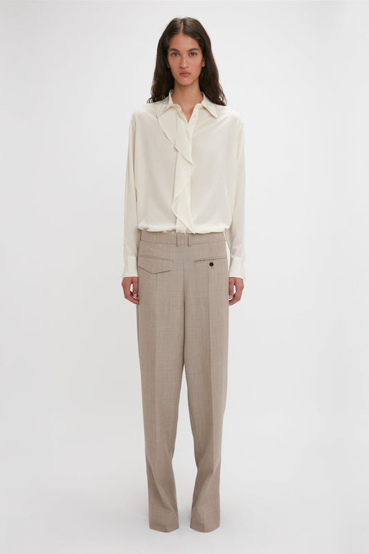 A person stands against a plain background wearing an Asymmetric Ruffle Blouse In Ivory by Victoria Beckham, featuring voluminous sleeves, and beige trousers.