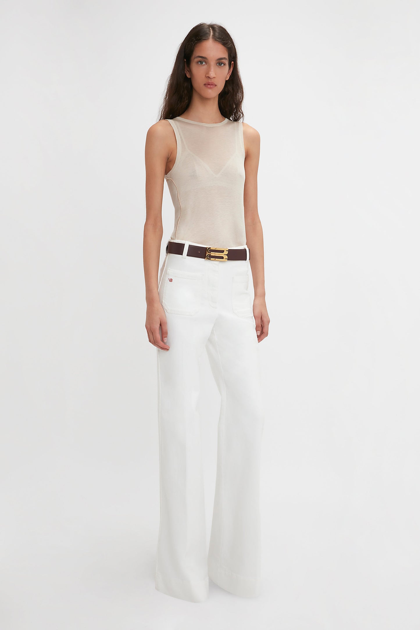 A woman with long dark hair is wearing a Victoria Beckham Lightweight Tank Top In Birch, white wide-leg pants, and a brown belt. She stands against a plain white background, reminiscent of the simplicity and elegance of birch trees.