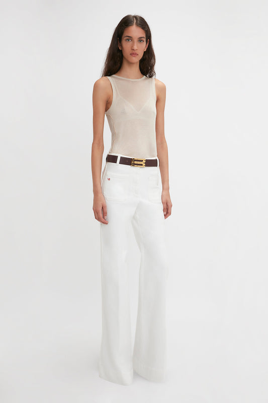 A woman with long dark hair is wearing a Victoria Beckham Lightweight Tank Top In Birch, white wide-leg pants, and a brown belt. She stands against a plain white background, reminiscent of the simplicity and elegance of birch trees.