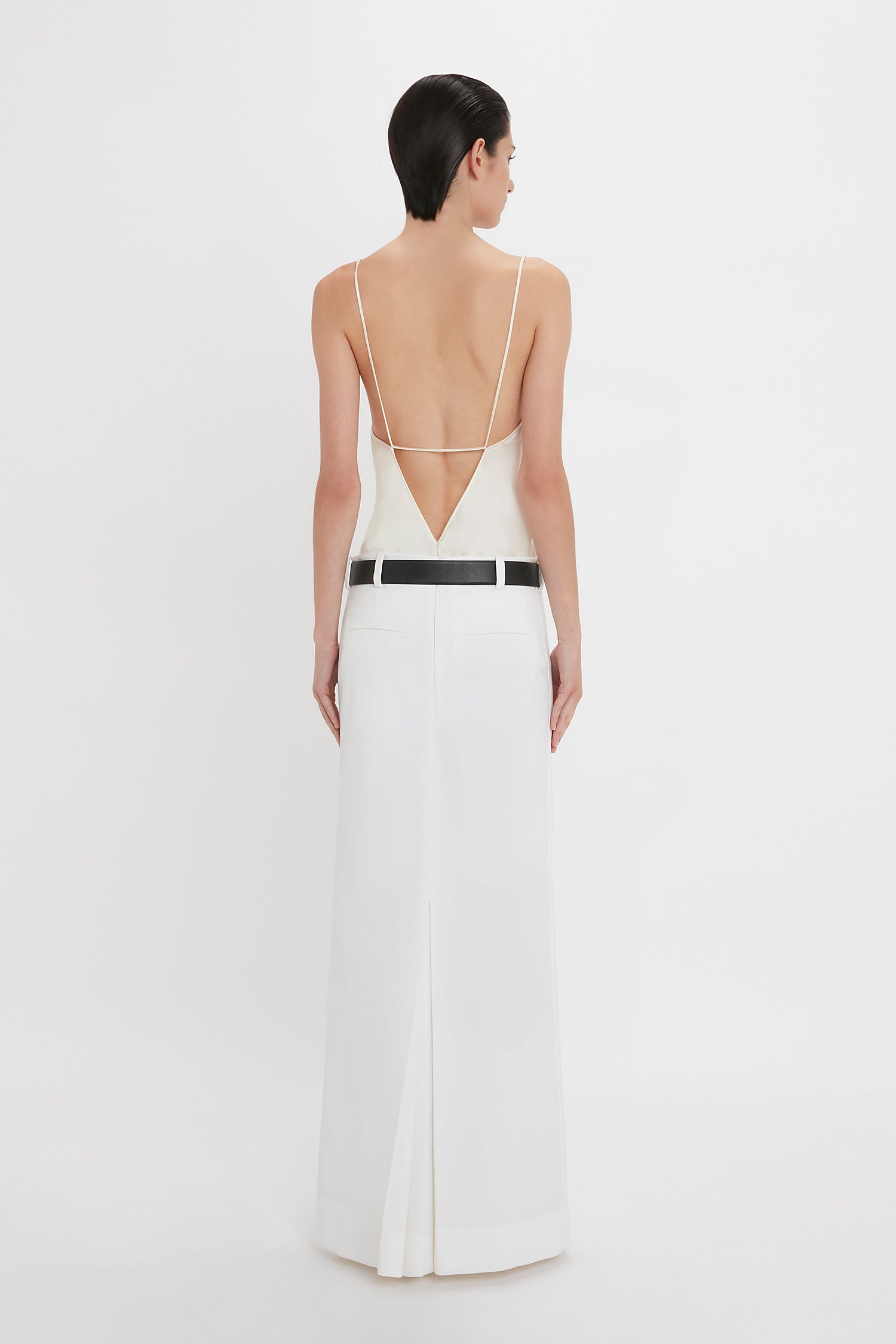 A person with short dark hair is wearing a backless white dress with a thin black belt and an Exclusive Tailored Floor-Length Pleated Skirt In Ivory by Victoria Beckham, standing and facing away from the camera against a plain white background.