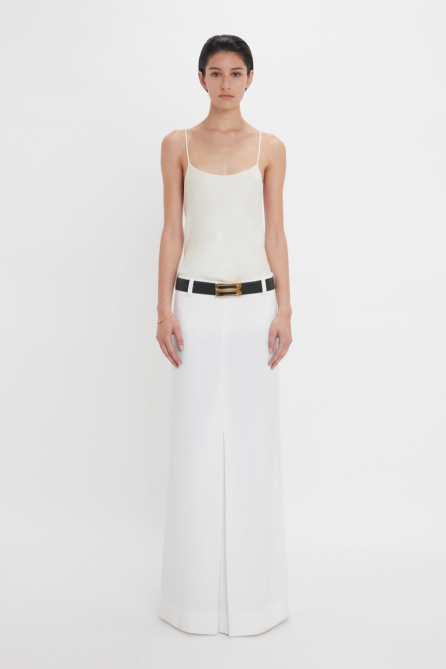 A person stands against a white background, wearing an Exclusive Cami Top In Ivory by Victoria Beckham and a long white skirt with a black belt featuring a gold buckle.