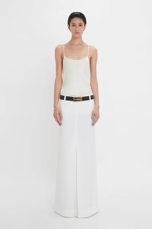 A person stands against a plain white background, wearing an ivory sleeveless top, an Exclusive Tailored Floor-Length Pleated Skirt In Ivory by Victoria Beckham, and a black belt with a gold buckle.