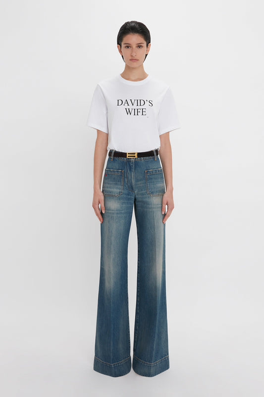 Person wearing a white T-shirt with "DAVID'S WIFE" written on it, paired with Victoria Beckham's Alina High Waisted Jean In Indigrey Wash and a black belt.