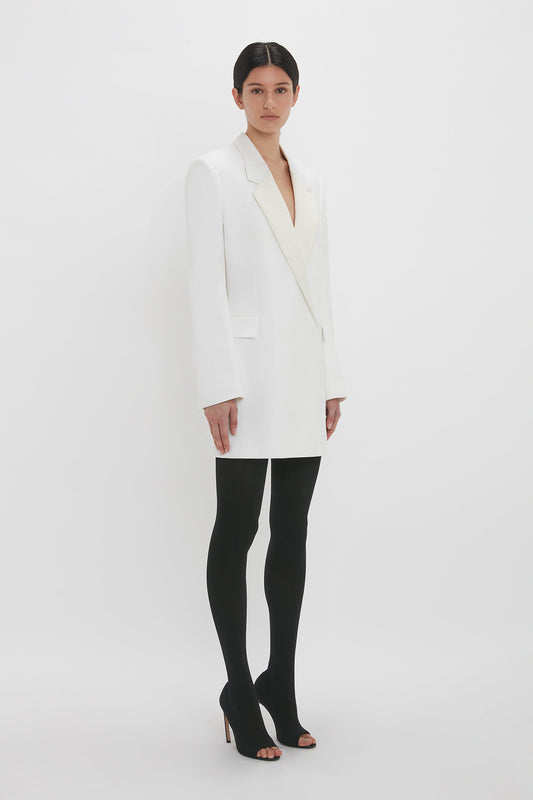 A woman stands against a white background, wearing a Victoria Beckham Exclusive Fold Shoulder Detail Dress In Ivory, black tights, and black high heels, looking directly at the camera.