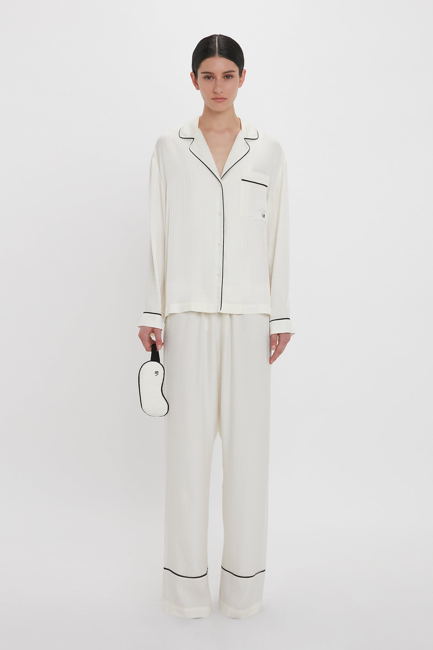 A woman stands against a white background, adorned in a luxurious 100% silk VB Monogram Pyjama Sleep Set In Ivory by Victoria Beckham with black piping. She holds a white sleep mask with a black border in her left hand, epitomizing elegant loungewear perfection.
