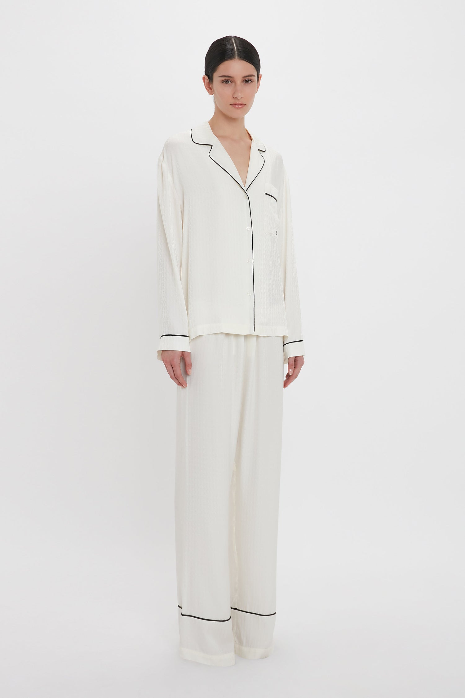 A person stands wearing a Victoria Beckham VB Monogram Pyjama Sleep Set In Ivory. The loungewear comprises a long-sleeve button-up top and matching wide-leg pants. The background is plain white.