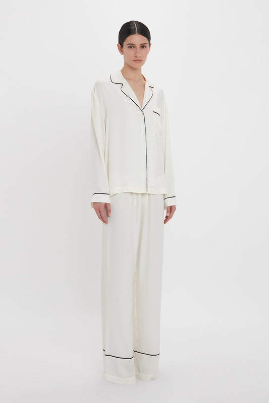 A person stands wearing a Victoria Beckham VB Monogram Pyjama Sleep Set In Ivory. The loungewear comprises a long-sleeve button-up top and matching wide-leg pants. The background is plain white.