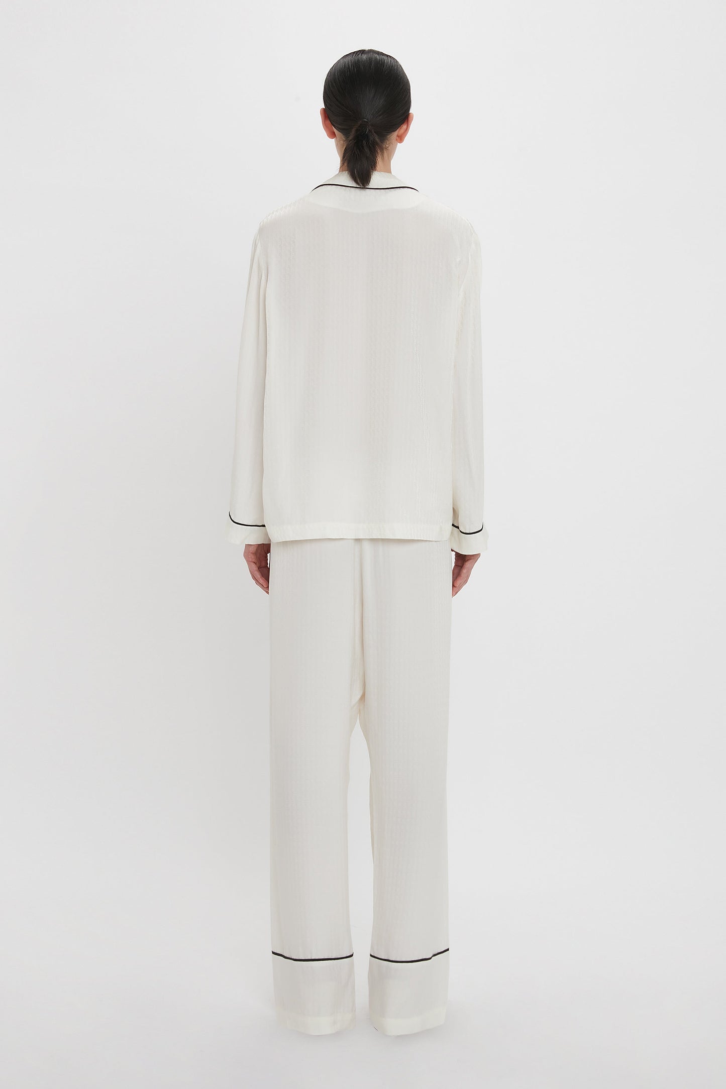 A person with dark hair is standing facing away, wearing a Victoria Beckham VB Monogram Pyjama Sleep Set in Ivory with black trim, against a plain white background.