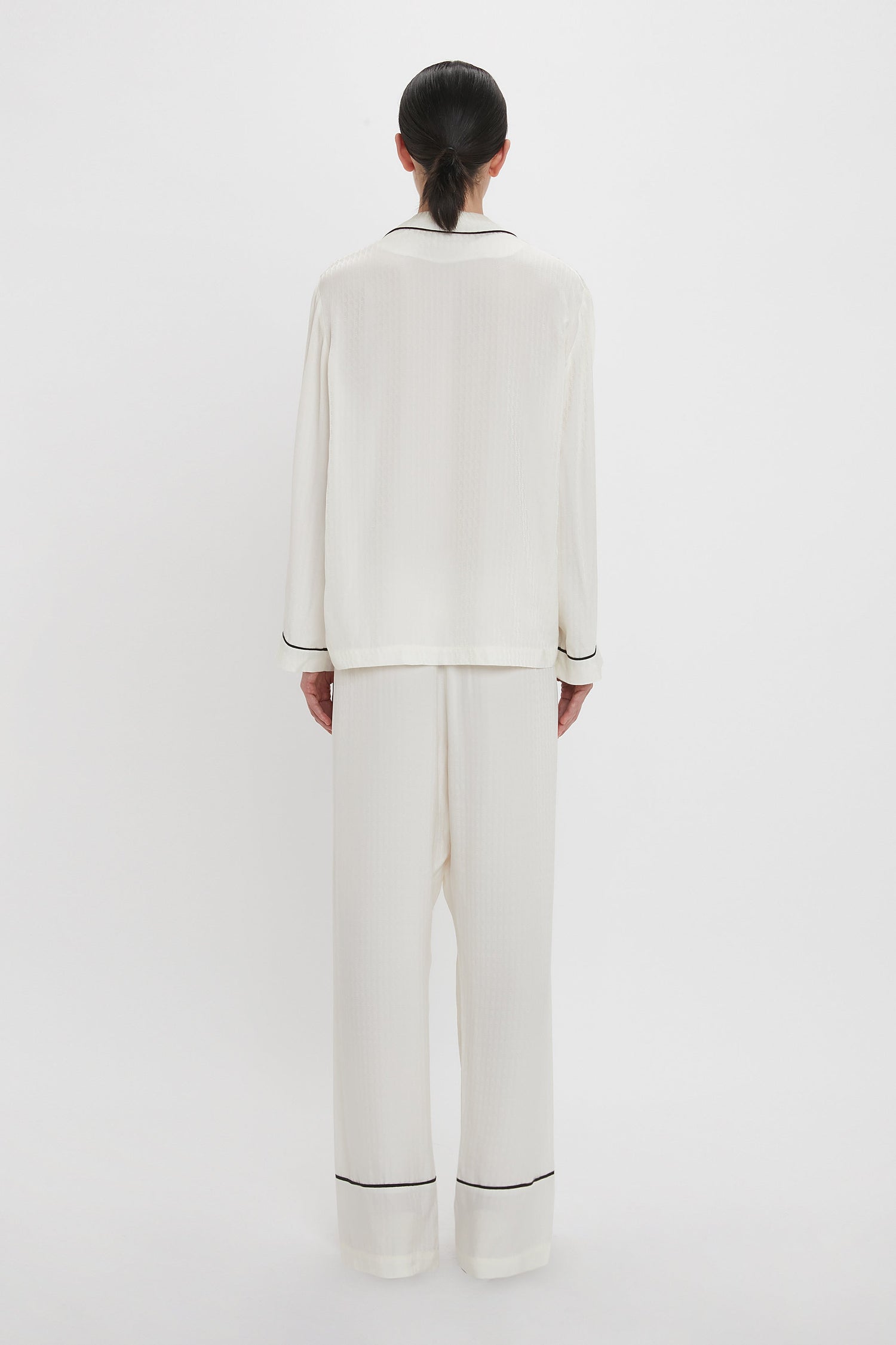 A person with dark hair is standing facing away, wearing a Victoria Beckham VB Monogram Pyjama Sleep Set in Ivory with black trim, against a plain white background.