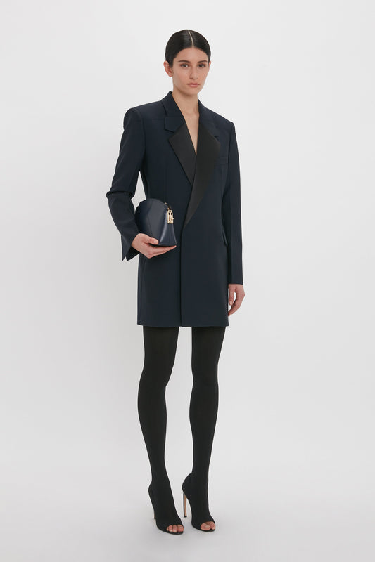 A person stands against a white background wearing the Victoria Beckham Exclusive Fold Shoulder Detail Dress In Midnight, black tights, and black open-toe heels, holding a small black clutch with a gold clasp.