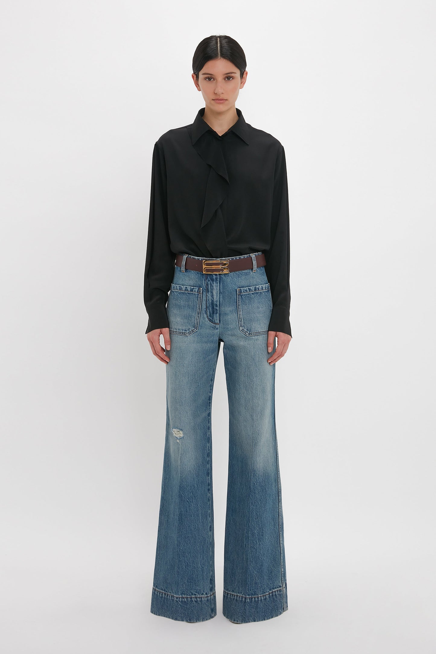 Person wearing a black blouse with a bow-tie collar, paired with high-waisted distressed Alina High Waisted Jean In Worn Blue Wash by Victoria Beckham and a brown belt, standing against a plain white background.