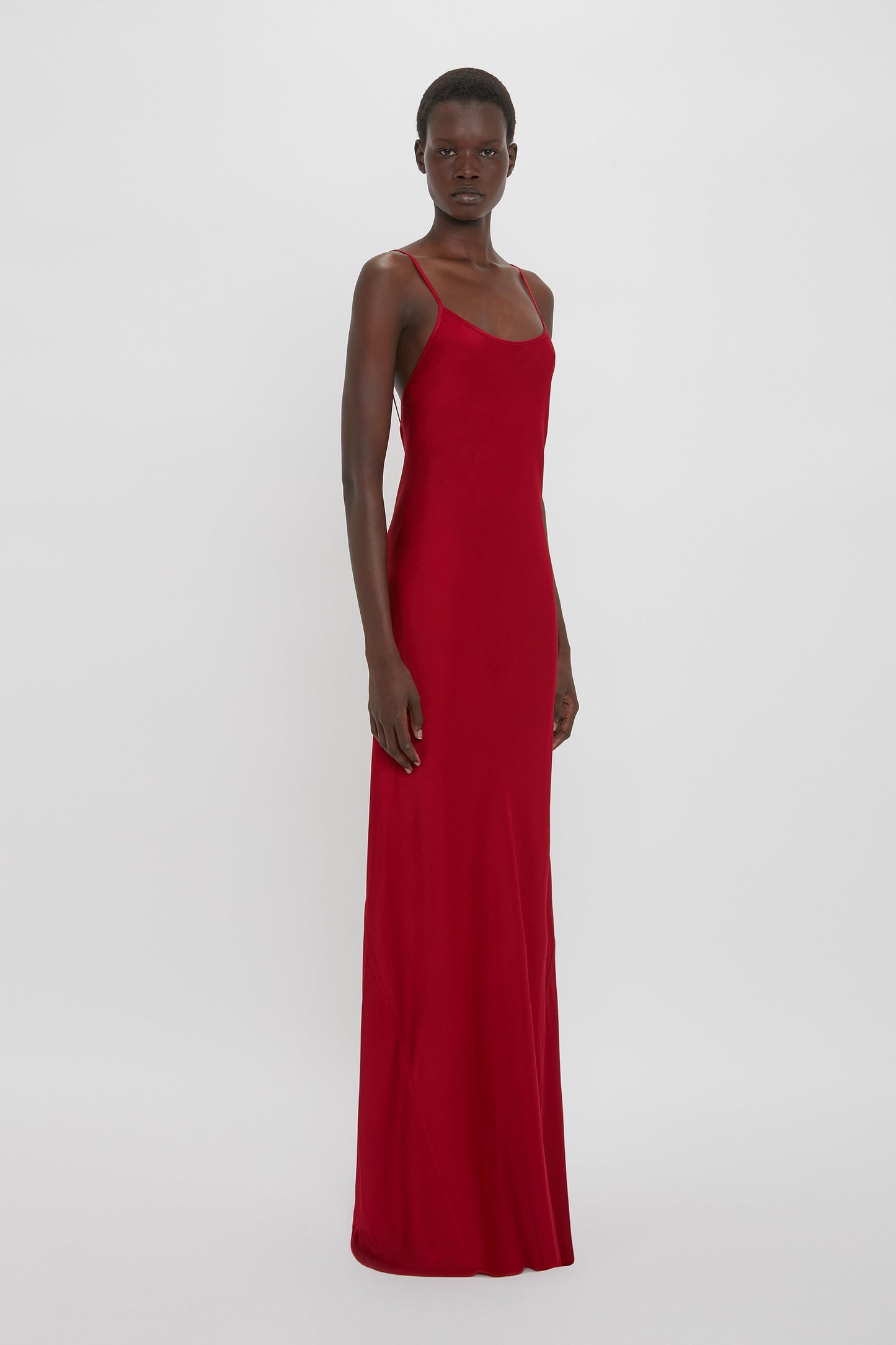 A person stands wearing a 1990s-inspired Victoria Beckham Low Back Cami Floor-Length Dress in Poppy Red, bias cut to perfection, against a plain white background.