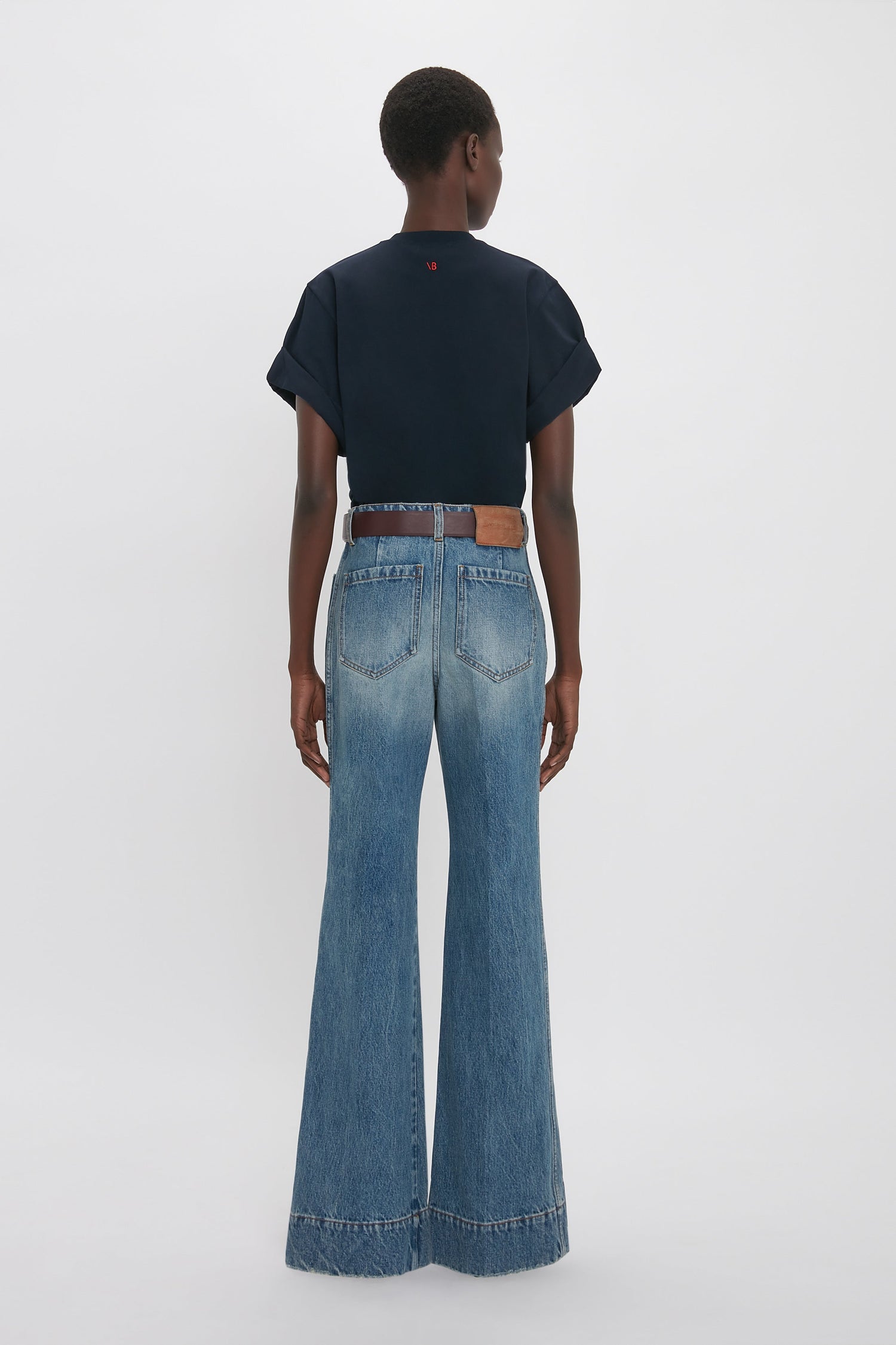A person stands facing away, wearing an oversized Victoria Beckham Asymmetric Relaxed Fit T-Shirt In Navy with asymmetric folded sleeves, paired with high-waisted, wide-leg blue denim jeans featuring visible back pockets.