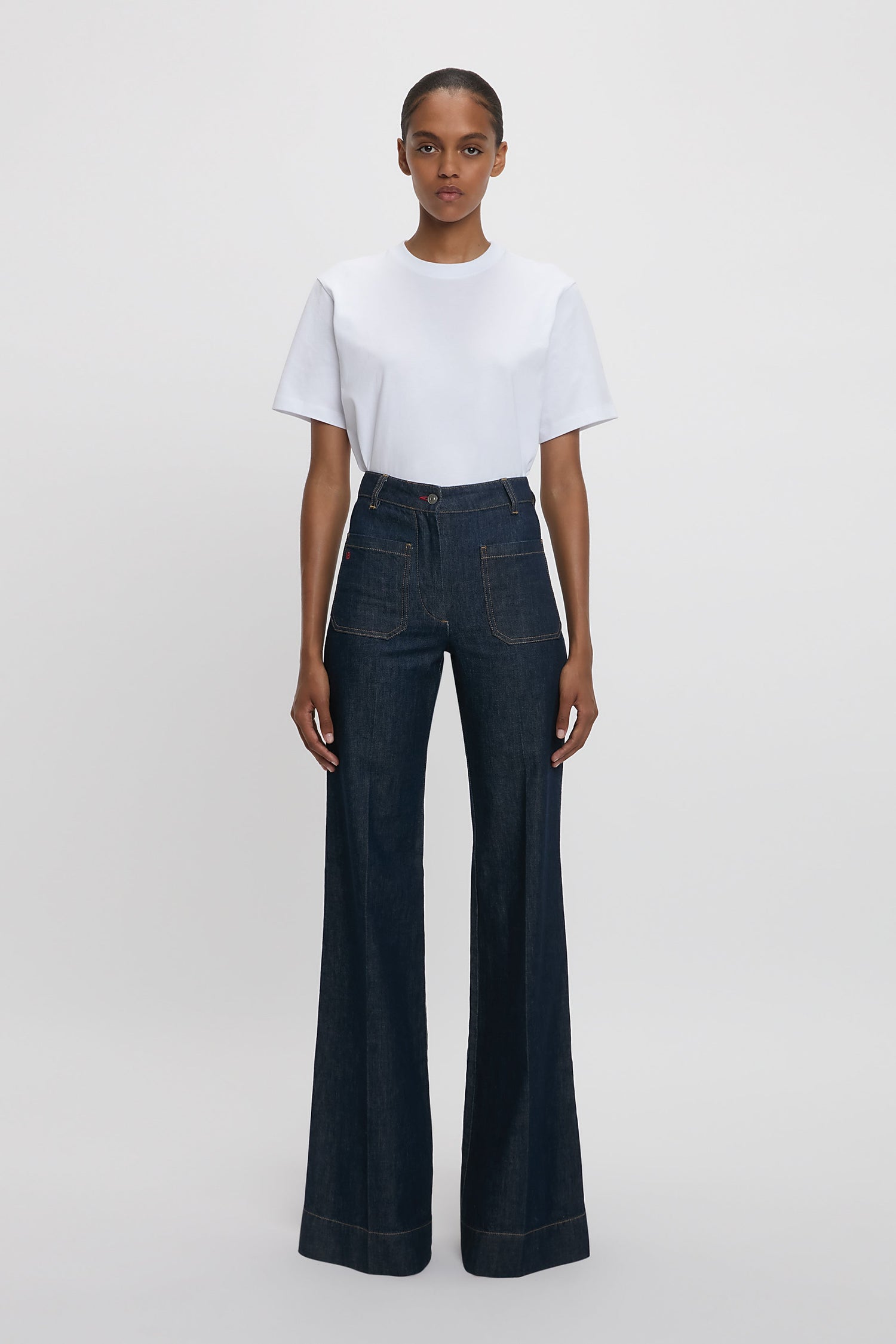 A woman standing against a white background wearing a white t-shirt and high-waisted, flared, indigo blue denim jeans with a branded leather patch at the waistband.