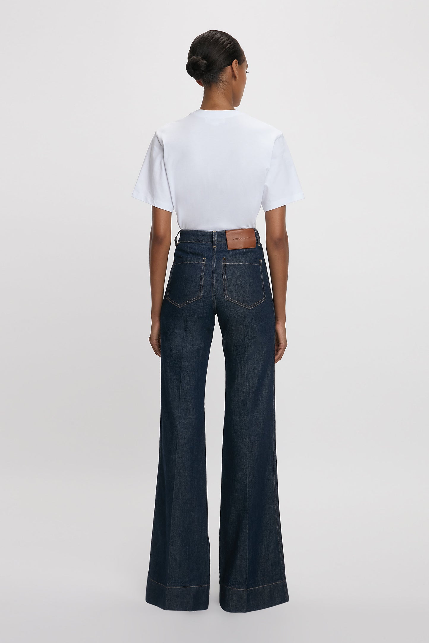 A woman standing towards a white background wearing a white t-shirt and high-waisted, flared, indigo blue denim jeans with a branded leather patch at the waistband.