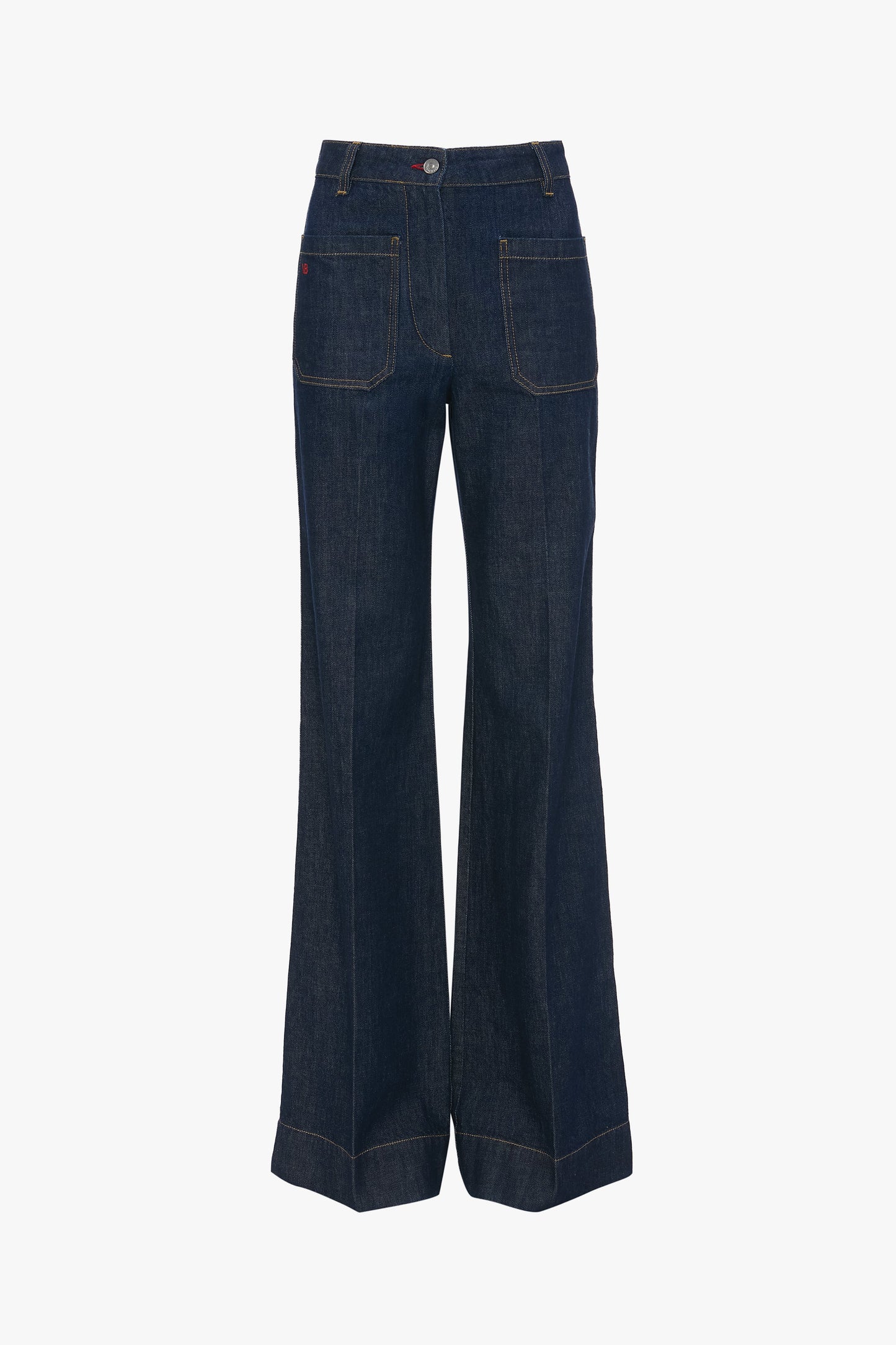 A woman standing against a white background wearing a white t-shirt and high-waisted, flared, indigo blue denim jeans with a branded leather patch at the waistband.